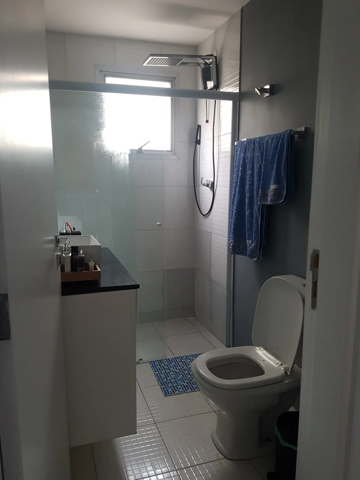 Banheiro antigo com box de vidro, azulejo branco, bancada de granito preto, vaso sanitário e toalha azul, antes da reforma