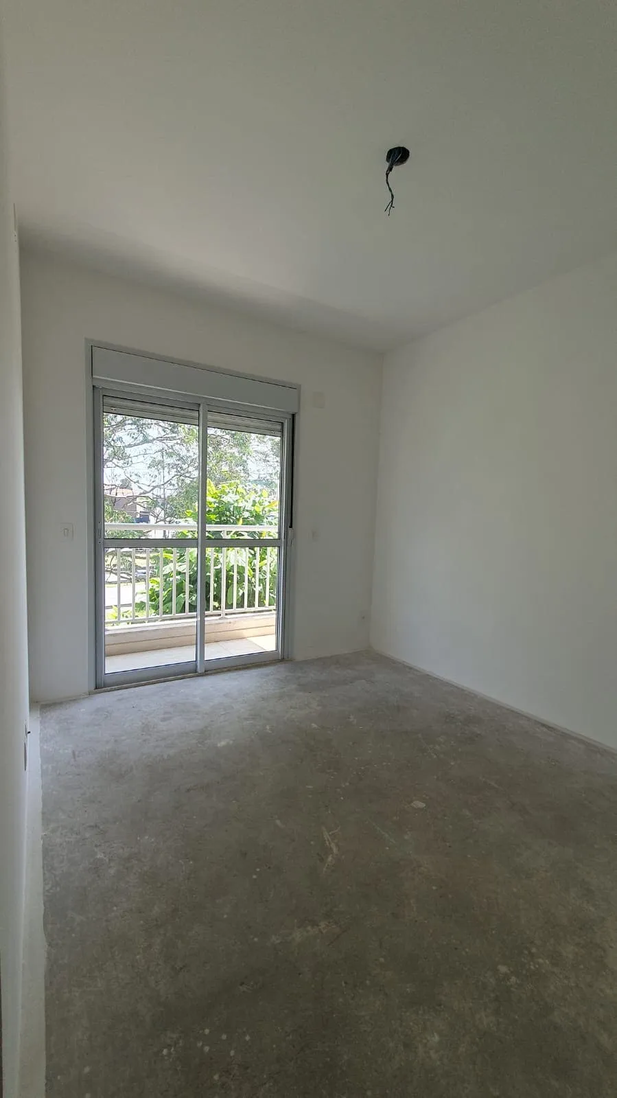 Quarto vazio com contrapiso cinza e porta de correr para varanda com vista verde, antes da reforma
