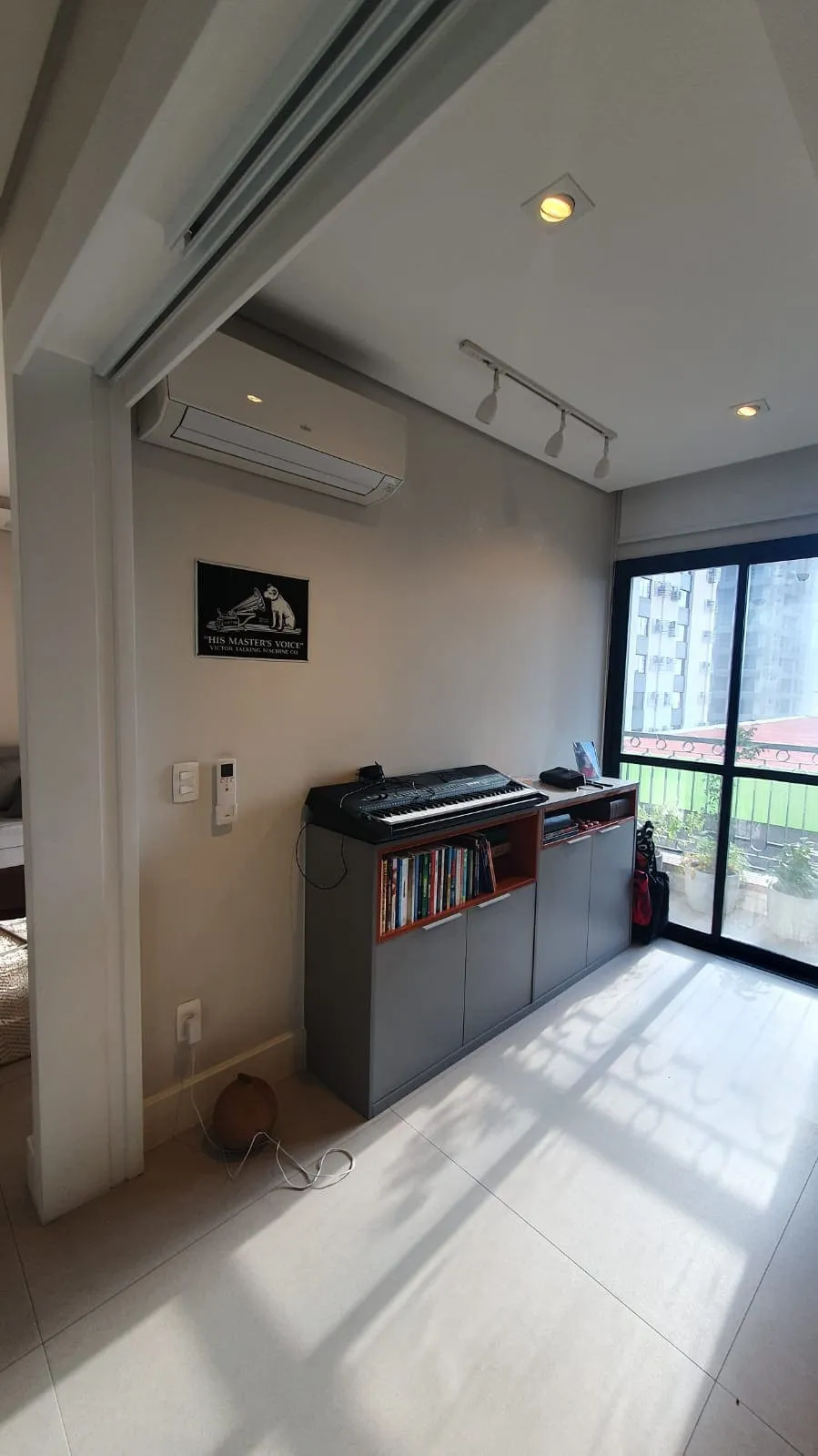 Sala antiga com balcão cinza, teclado musical, pôster emoldurado e porta de vidro para a varanda, antes da reforma
