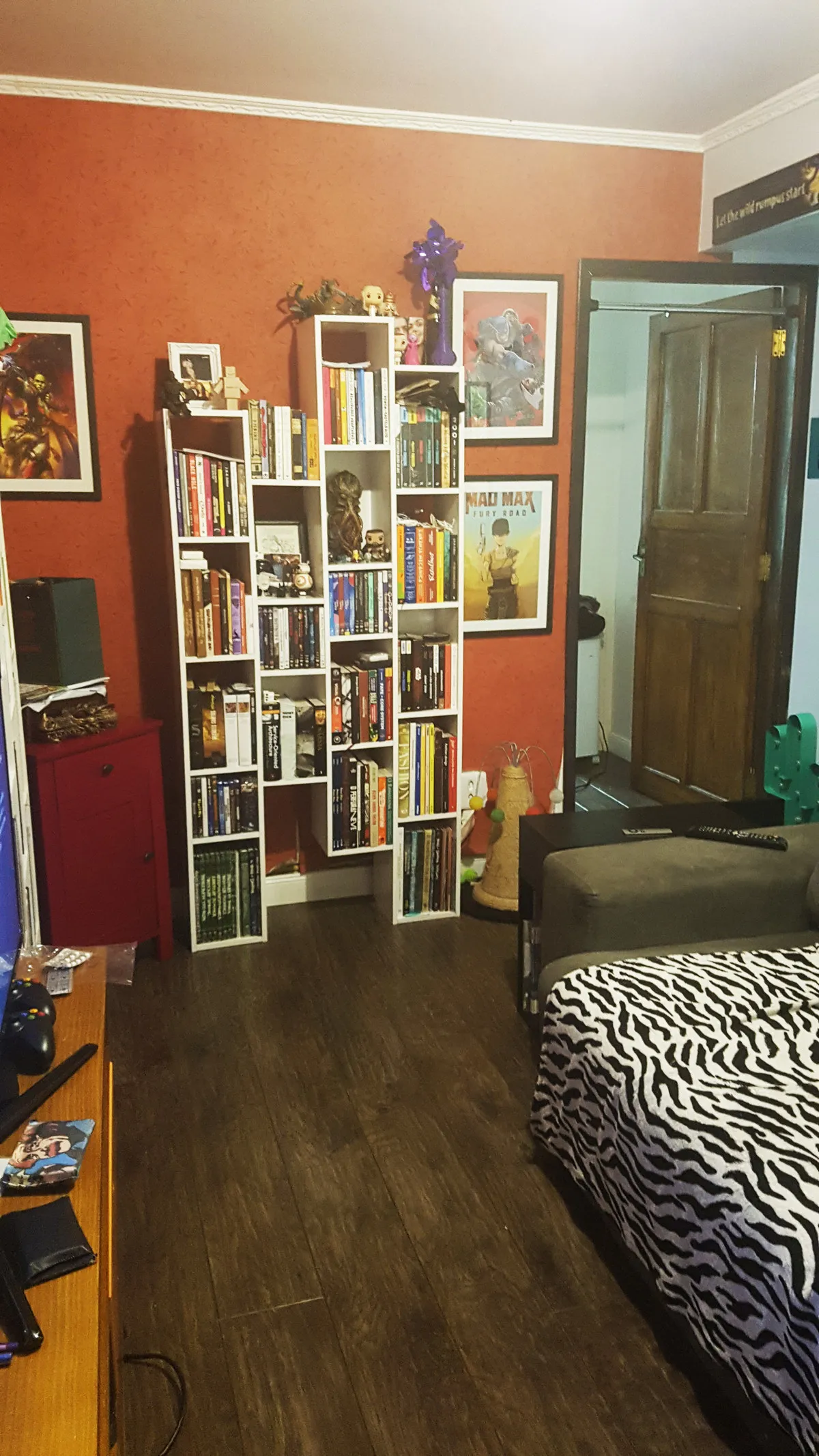 Sala com paredes alaranjadas, estantes brancas cheias de livros e colecionáveis, pôster do Mad Max e piso de madeira escura, antes da reforma