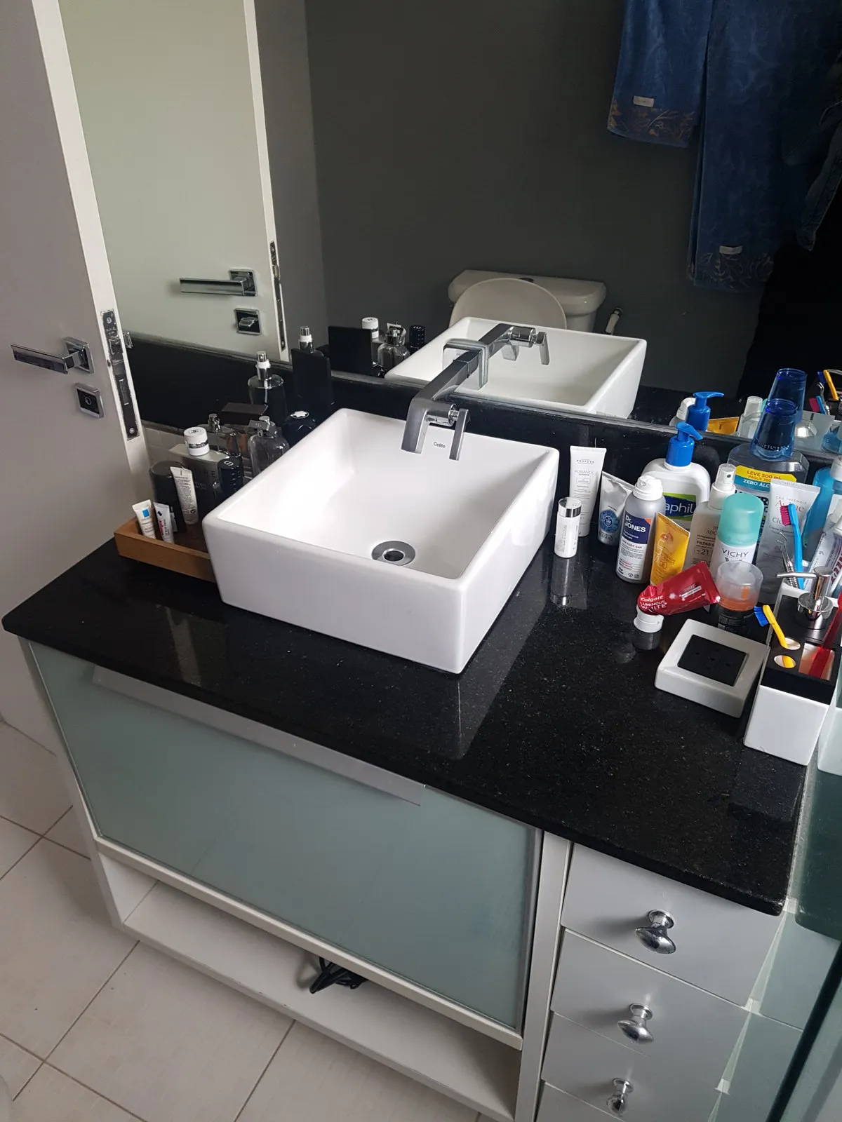 Bancada de banheiro em granito preto com cuba de apoio branca e produtos espalhados diante do espelho, antes da reforma
