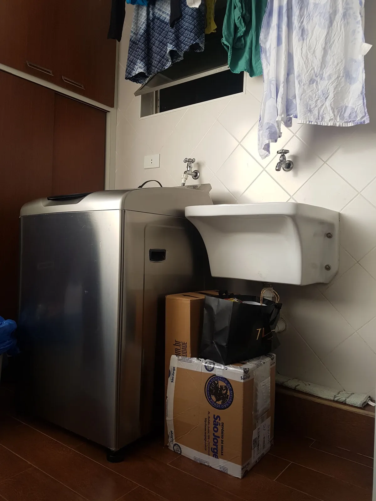 Área de serviço com tanque branco, máquina de lavar prateada e caixas de papelão no chão, antes da reforma