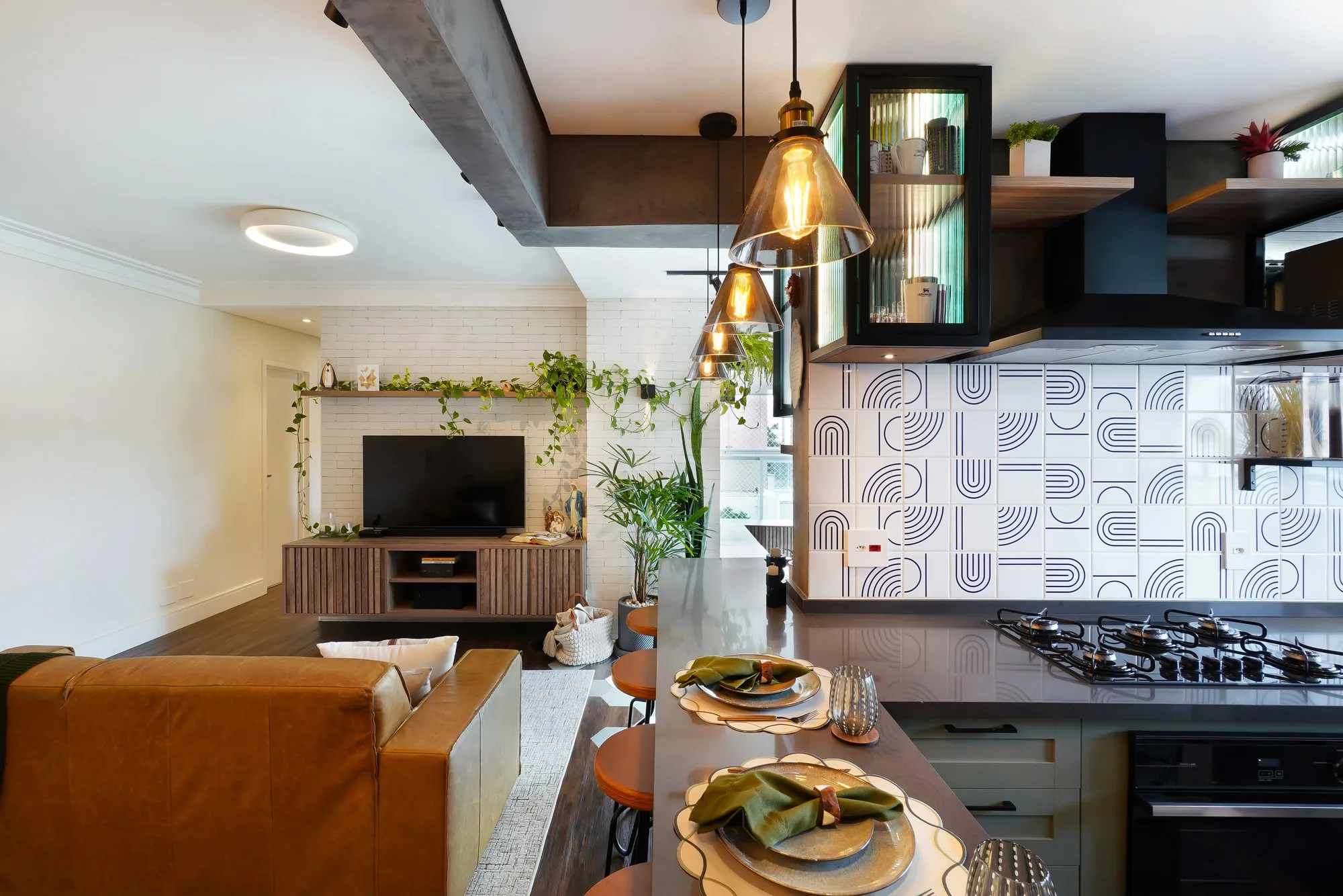 Sala de estar integrada à cozinha, com sofá de couro caramelo, parede de tijolo branco com plantas, ilha com cooktop e mesa posta, sob pendentes de vidro