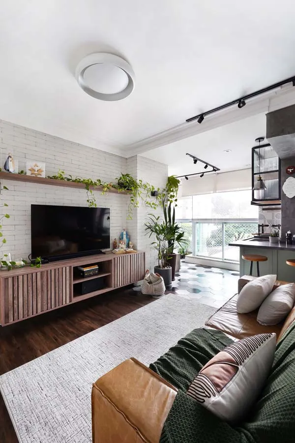 Sala de estar com sofá de couro caramelo e manta verde, painel de tijolo branco com prateleira de madeira e plantas, TV sobre rack ripado e piso de madeira escura