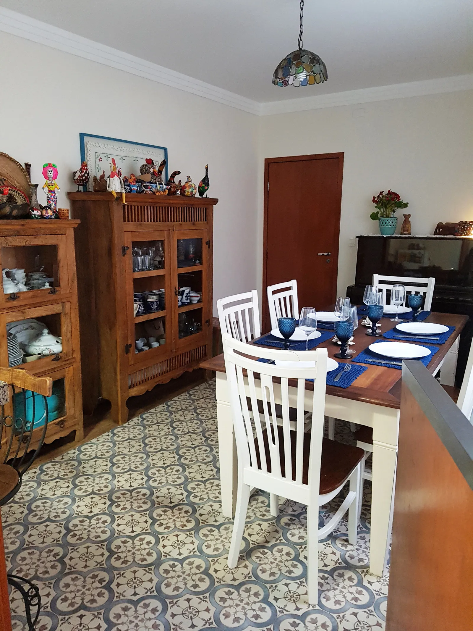Sala de jantar rústica com mesa de madeira posta, cadeiras brancas e piso de ladrilho hidráulico, com cristaleiras de madeira ao fundo