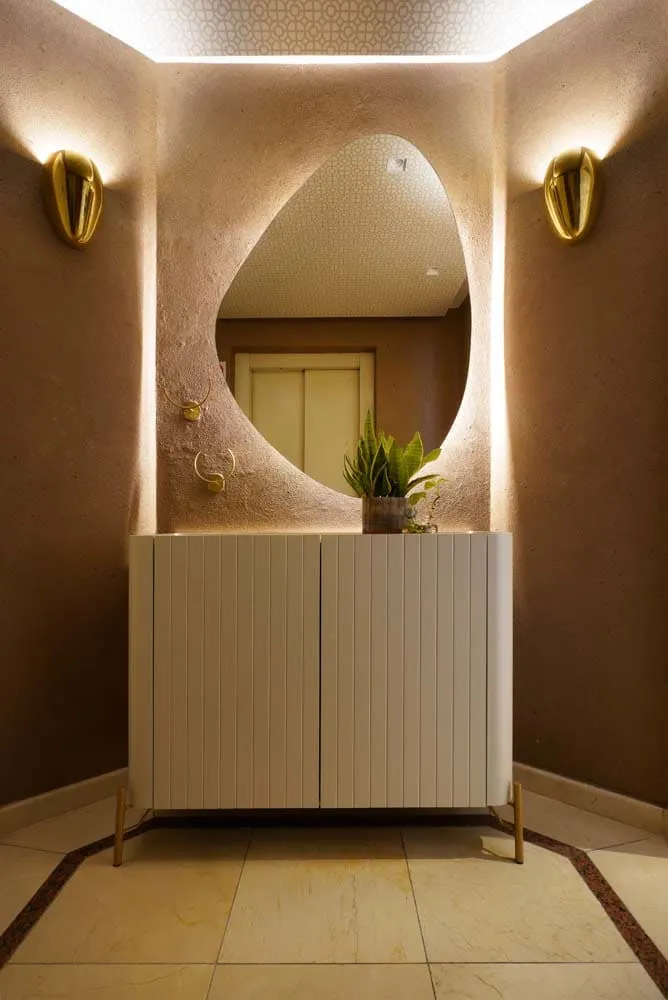 Lavabo com nicho de espelho orgânico, parede em textura terracota, arandelas douradas e aparador ripado branco sobre piso de mármore
