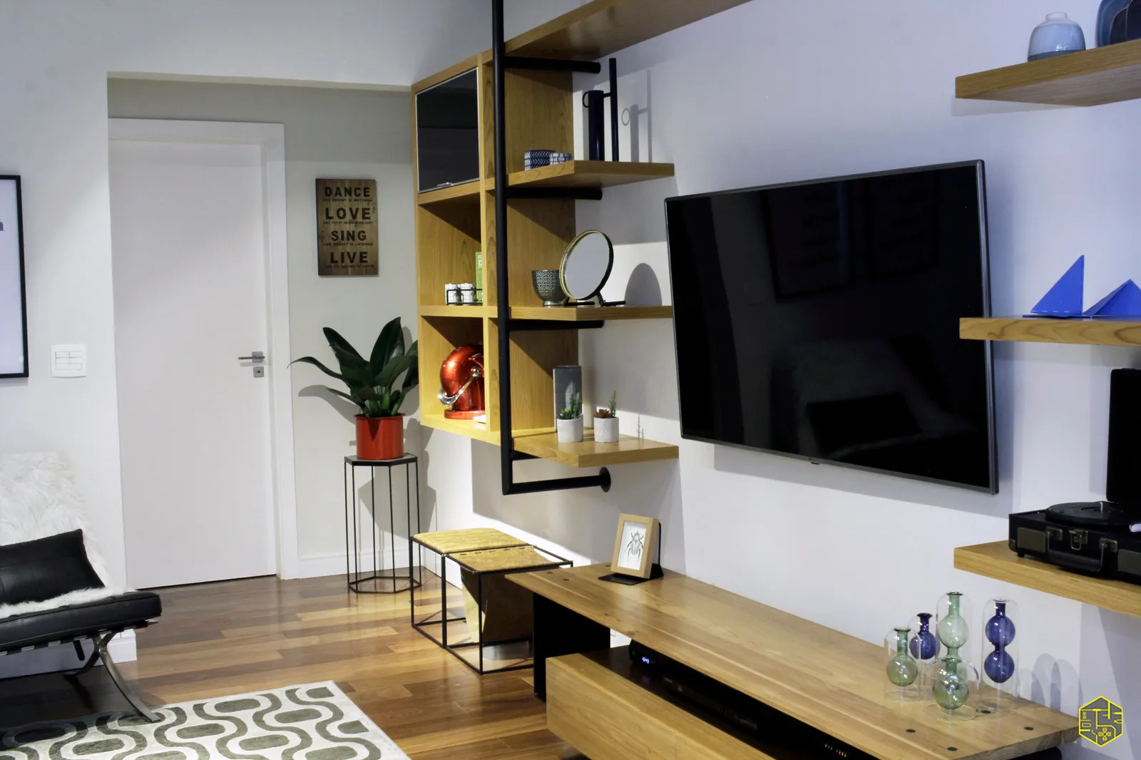 Sala com estante de aço tubular preto e prateleiras de madeira, TV na parede, poltrona Barcelona preta e piso de madeira