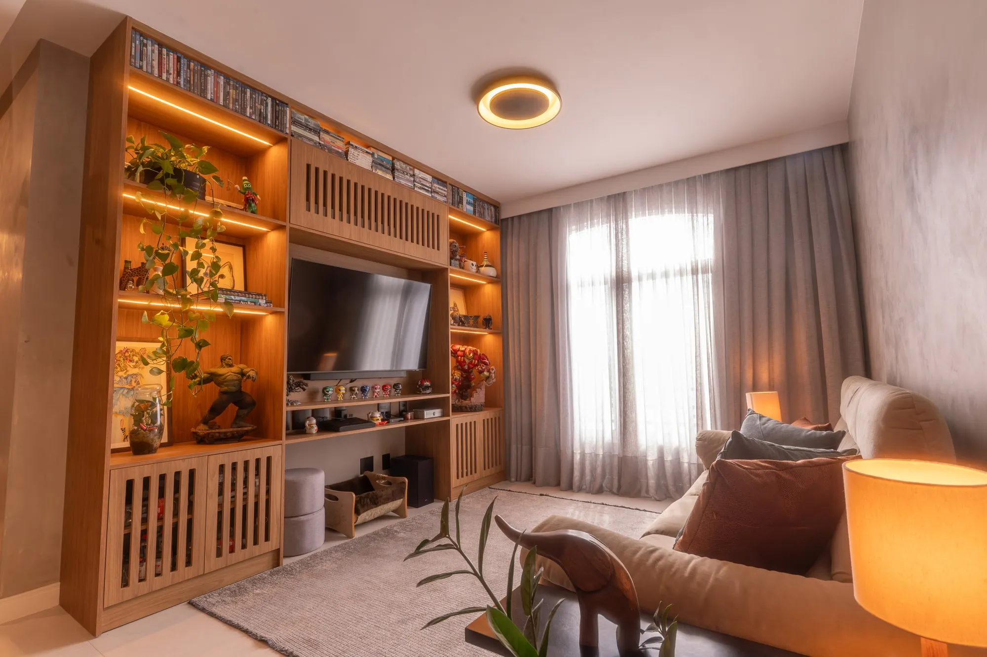Sala de estar com estante ripada de madeira iluminada, TV, colecionáveis, sofá bege e cortina cinza