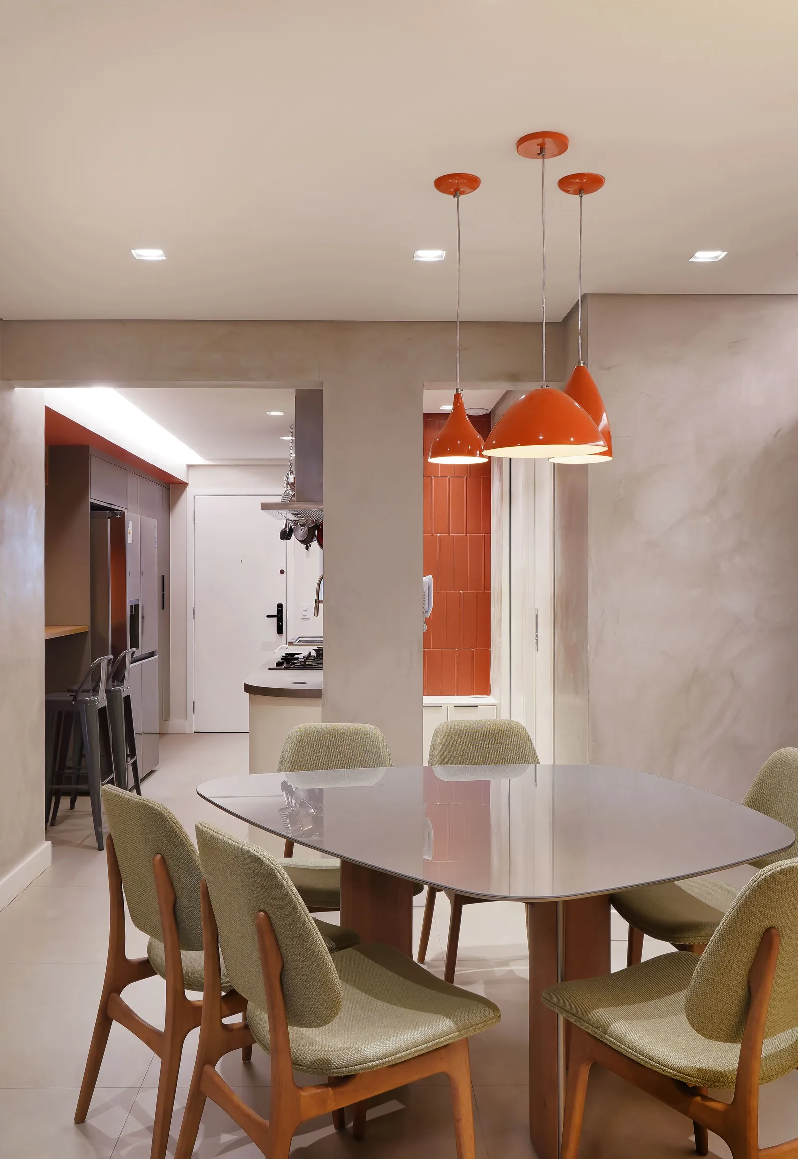 Sala de jantar com mesa de madeira e cadeiras verdes, pendentes laranja e vista para a cozinha ao fundo