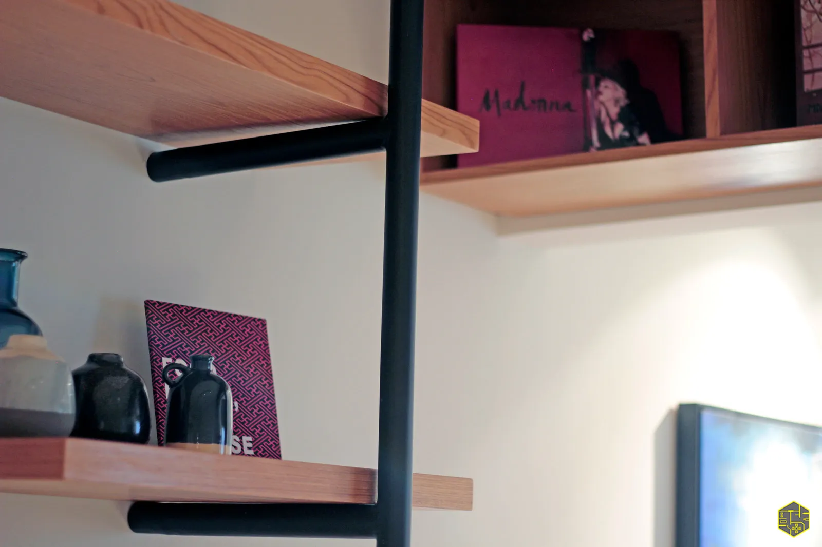 Detalhe das prateleiras de madeira com livro da Madonna, vasos escuros e a estrutura tubular preta em primeiro plano
