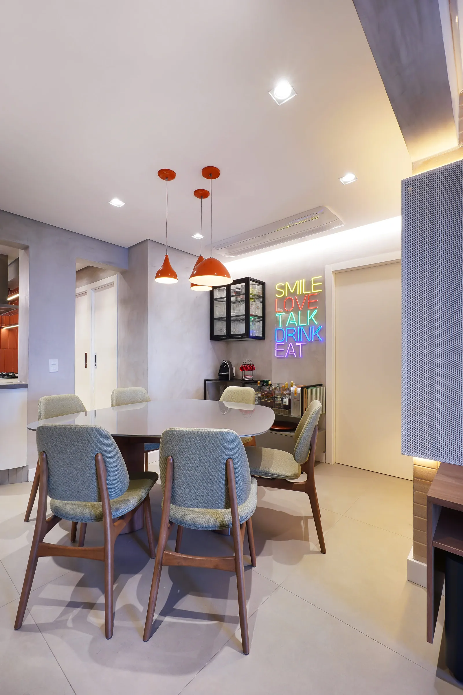 Sala de jantar com mesa oval, cadeiras verdes, pendentes laranja, cristaleira preta e letreiro de neon colorido