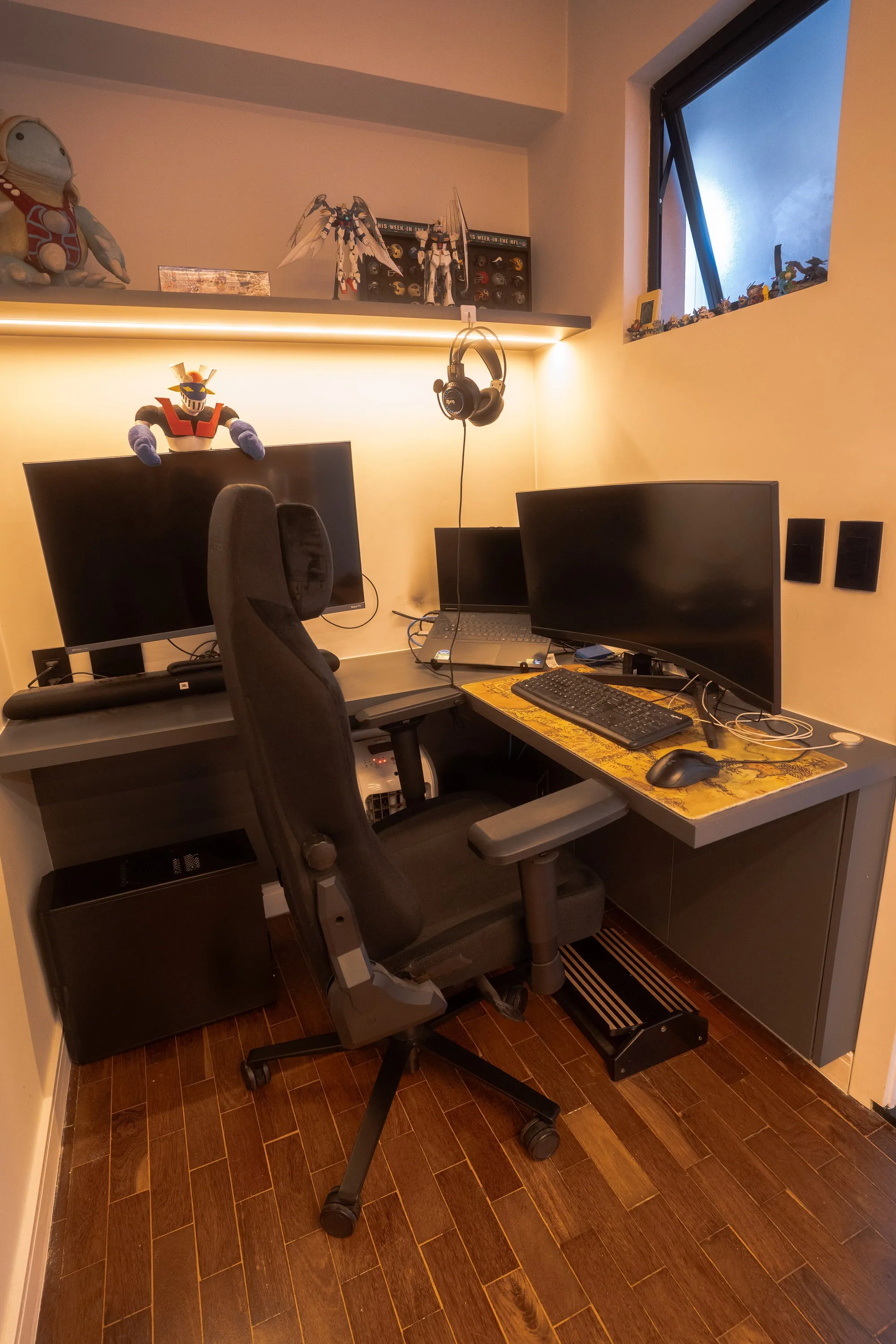 Estação de trabalho em L com dois monitores, cadeira gamer e prateleira de colecionáveis