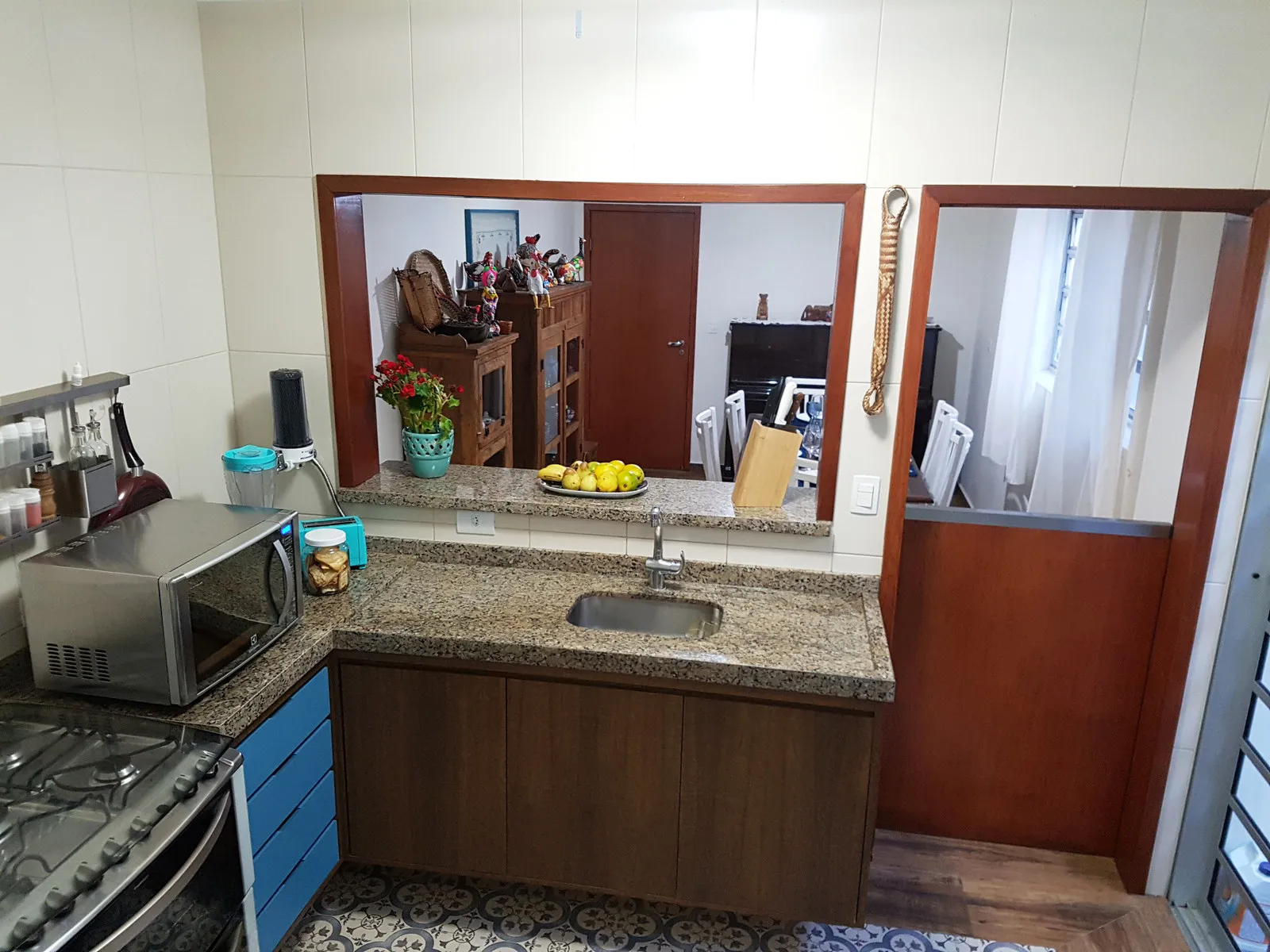 Cozinha integrada com bancada de granito, marcenaria amadeirada com gavetas azuis e o passa-prates aberto para a sala