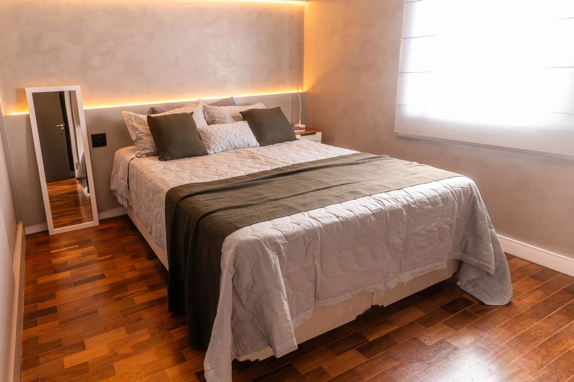 Dormitório com cama de casal, cabeceira de cimento queimado iluminada, roupa de cama verde musgo e espelho