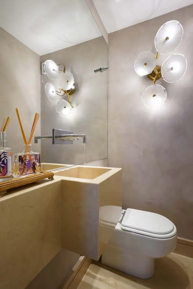 Lavabo com cuba esculpida em mármore bege, torneira de parede cromada e arandelas de vidro em flor sobre cimento queimado