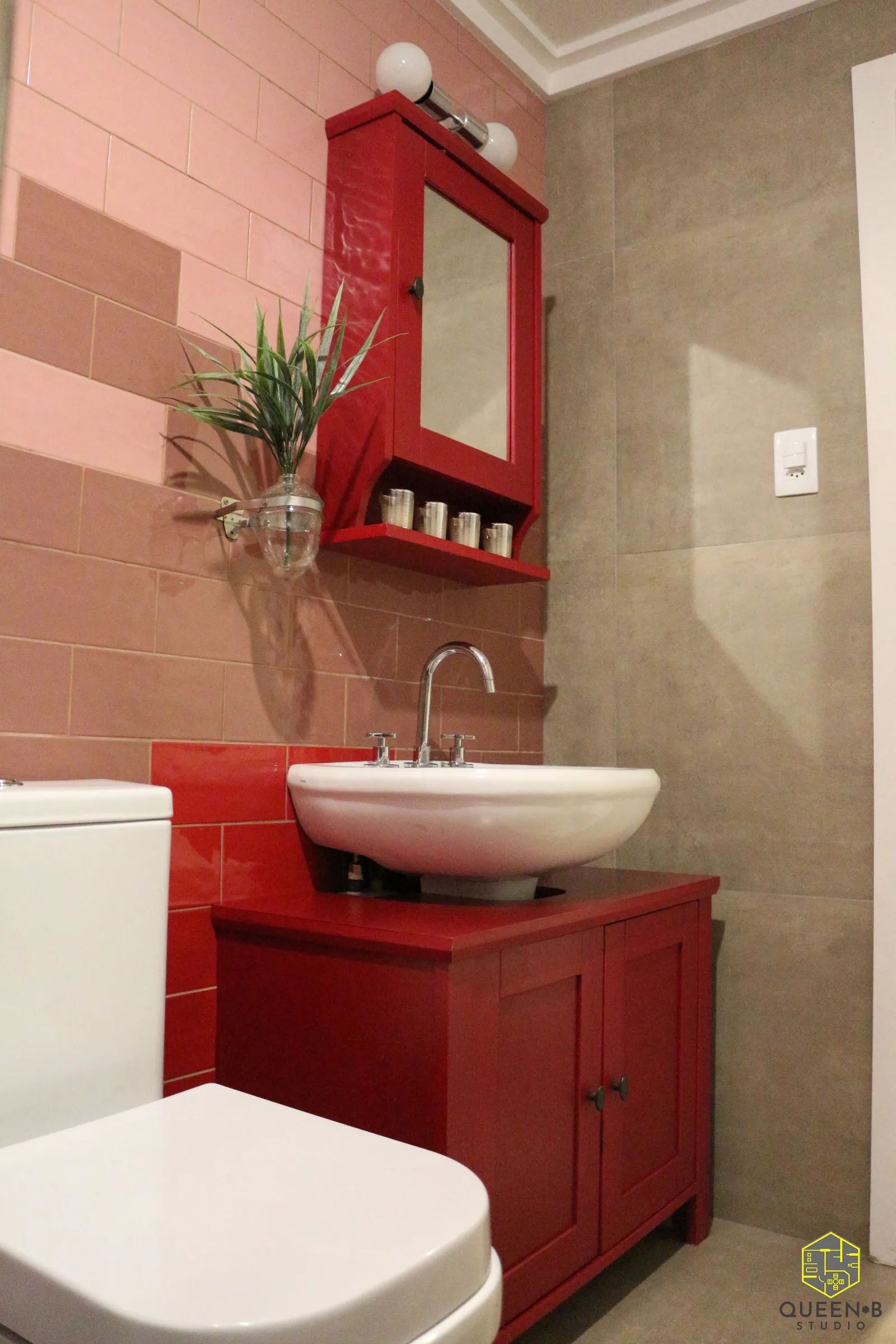 Banheiro com azulejo rosa e bege, gabinete e armário-espelho vermelhos, cuba de apoio branca e planta em vaso de vidro