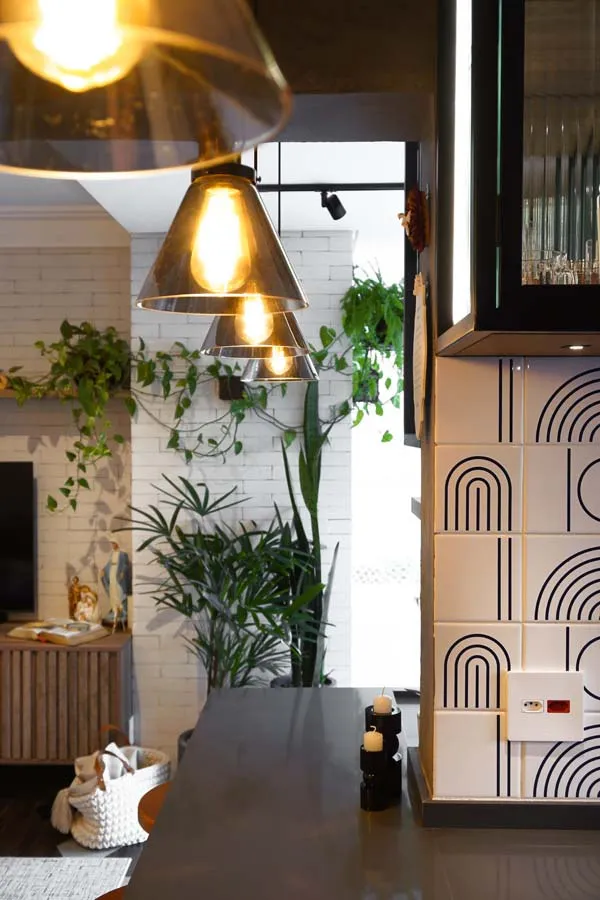 Detalhe dos pendentes de vidro sobre a península cinza, com painel de tijolo branco e plantas ao fundo e o revestimento geométrico à direita