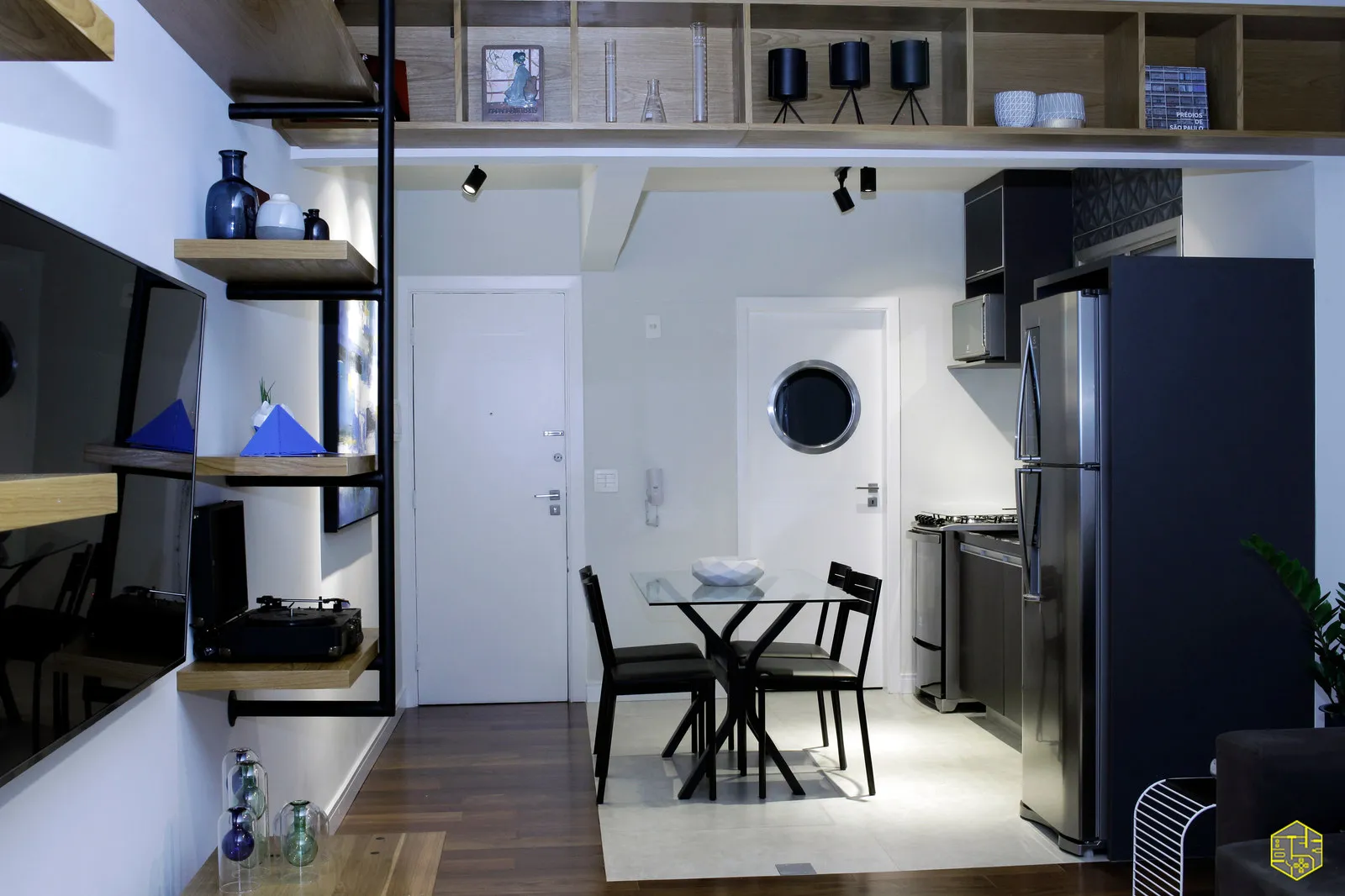 Sala e cozinha integradas com mesa de jantar de vidro e cadeiras pretas, geladeira inox e prateleiras de madeira com decoração