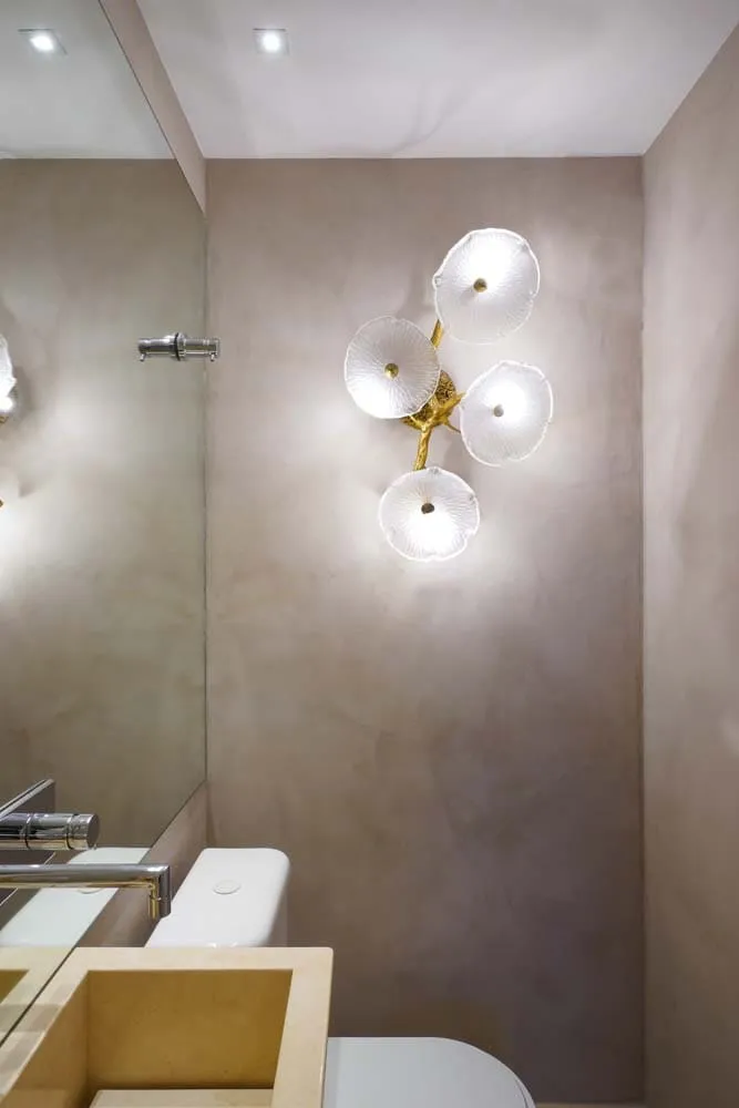Lavabo com arandela de pétalas de vidro e miolo dourado na parede de cimento queimado, com cuba de mármore bege