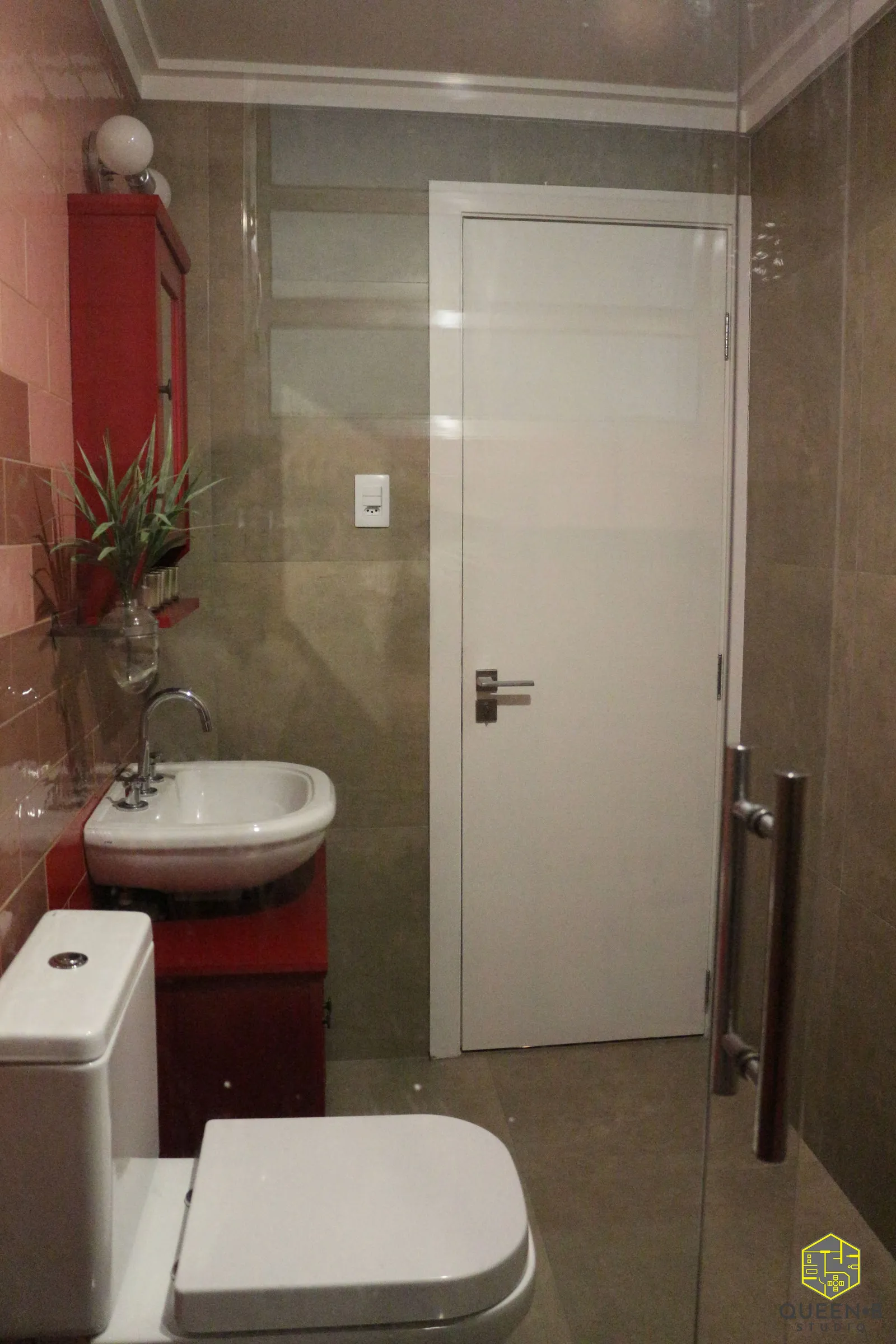 Banheiro com parede de porcelanato bege, porta branca, box de vidro e gabinete vermelho com cuba e vaso sanitário brancos