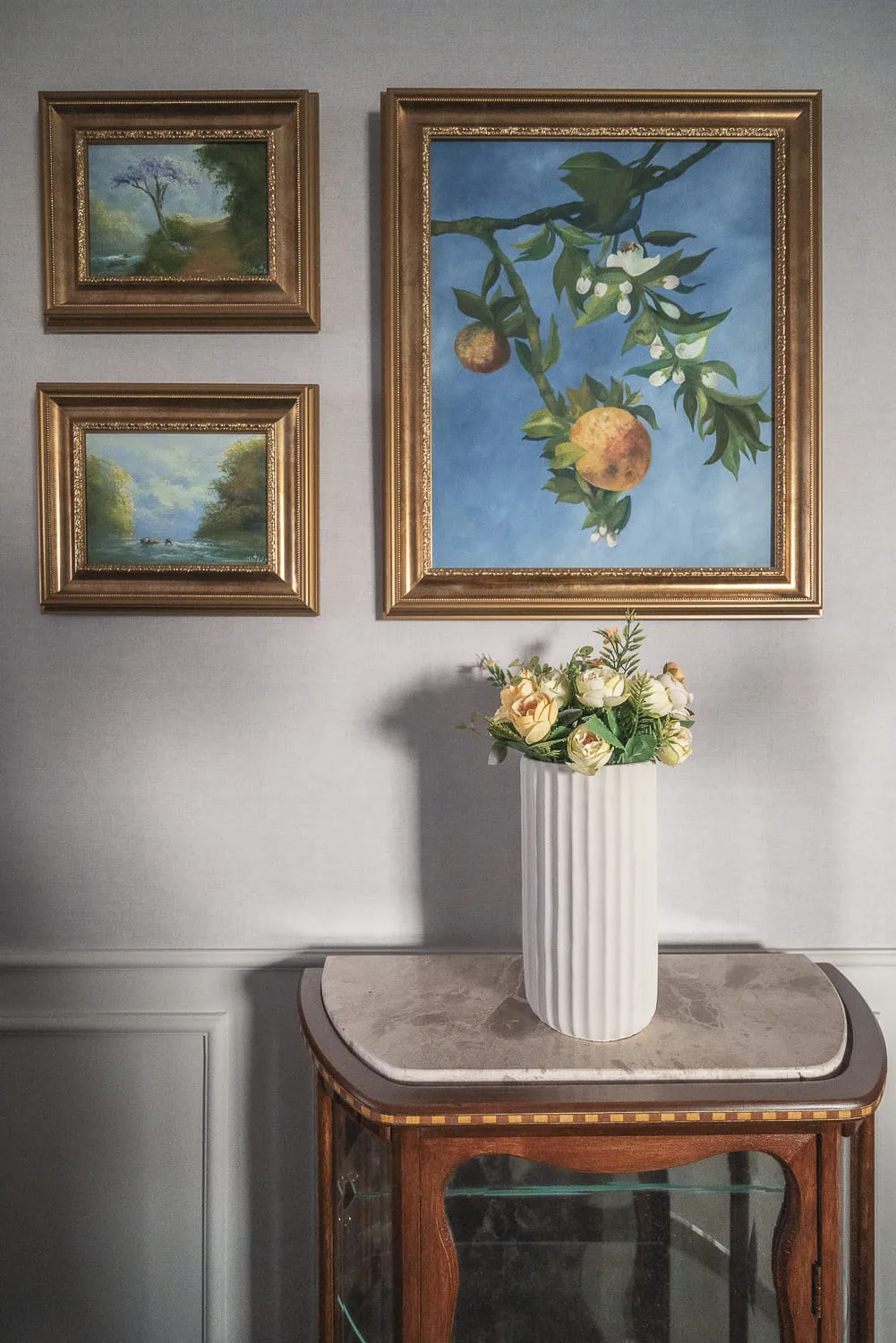 Detalhe de parede com quadros a óleo emoldurados em dourado e vaso branco de flores sobre cristaleira de madeira