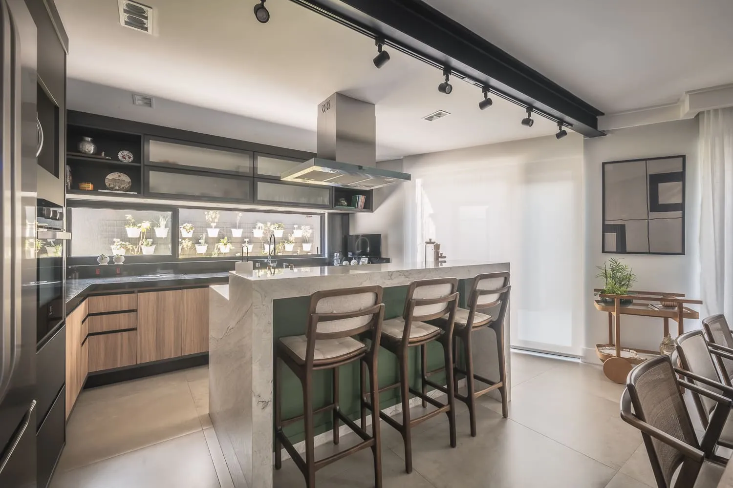 Cozinha com ilha de mármore, banquetas verdes e jardim de temperos iluminado na parede, aberta para a sala