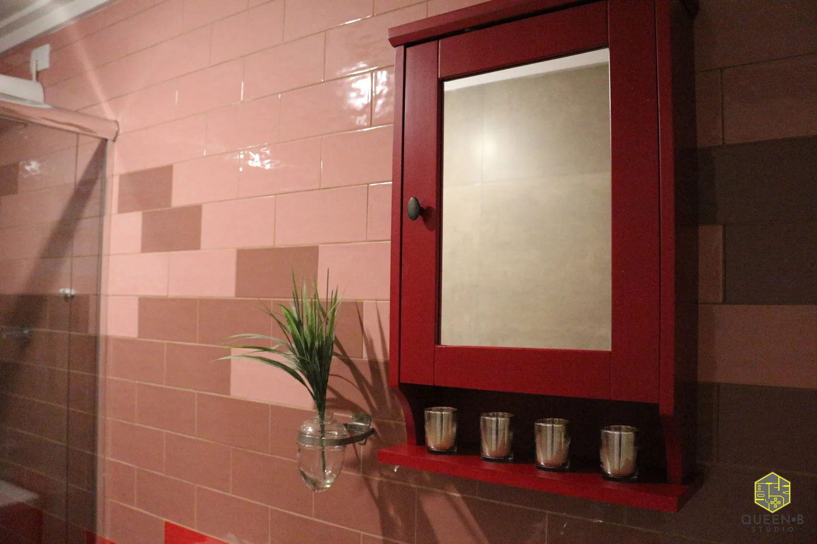 Detalhe do armário-espelho vermelho com copos metálicos na prateleira, parede de azulejo rosa e vaso de vidro com planta
