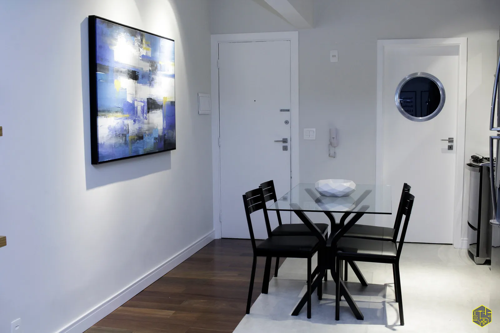 Sala de jantar com mesa de vidro e cadeiras pretas, quadro abstrato azul, porta branca e espelho redondo tipo escotilha