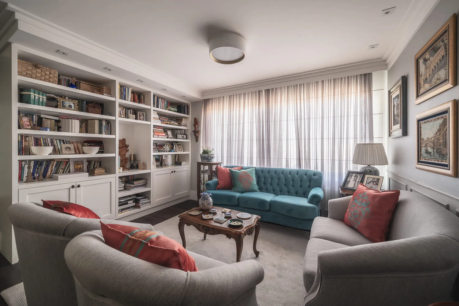 Sala de estar com sofá azul-petróleo capitonê, poltronas cinza, estante branca de livros e almofadas vermelhas