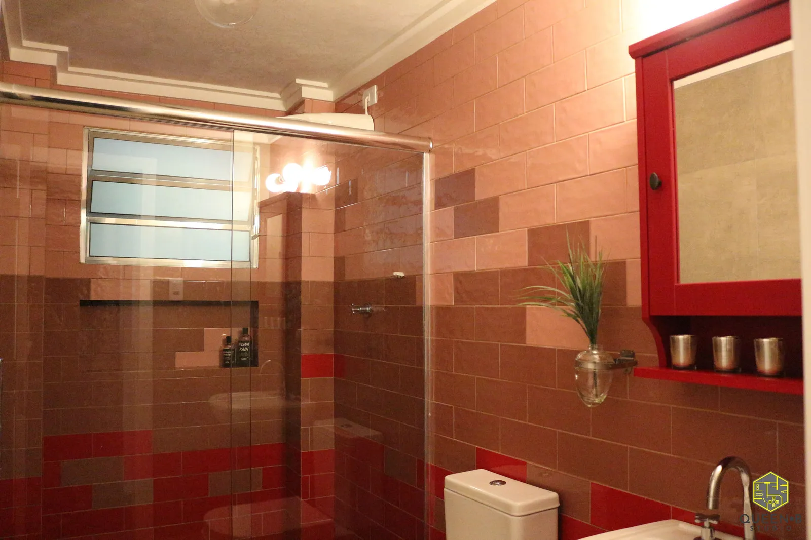 Banheiro com box de vidro, azulejos em tons de rosa, marrom e vermelho, janela basculante e armário-espelho vermelho