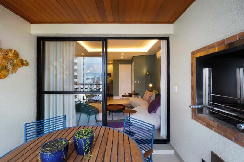 Varanda com mesa de madeira, cadeiras azuis e churrasqueira de tijolinho, integrada à sala pela porta de vidro