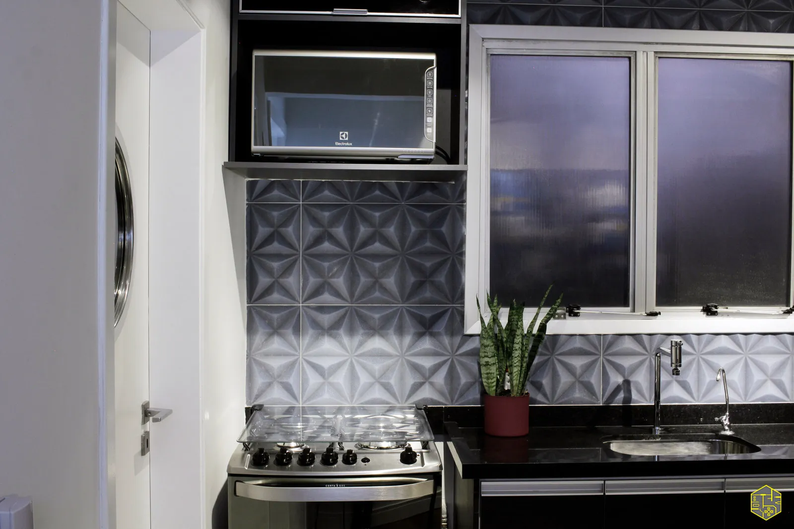 Detalhe da cozinha com azulejo geométrico 3D, fogão inox, micro-ondas embutido e bancada de granito preto com cuba