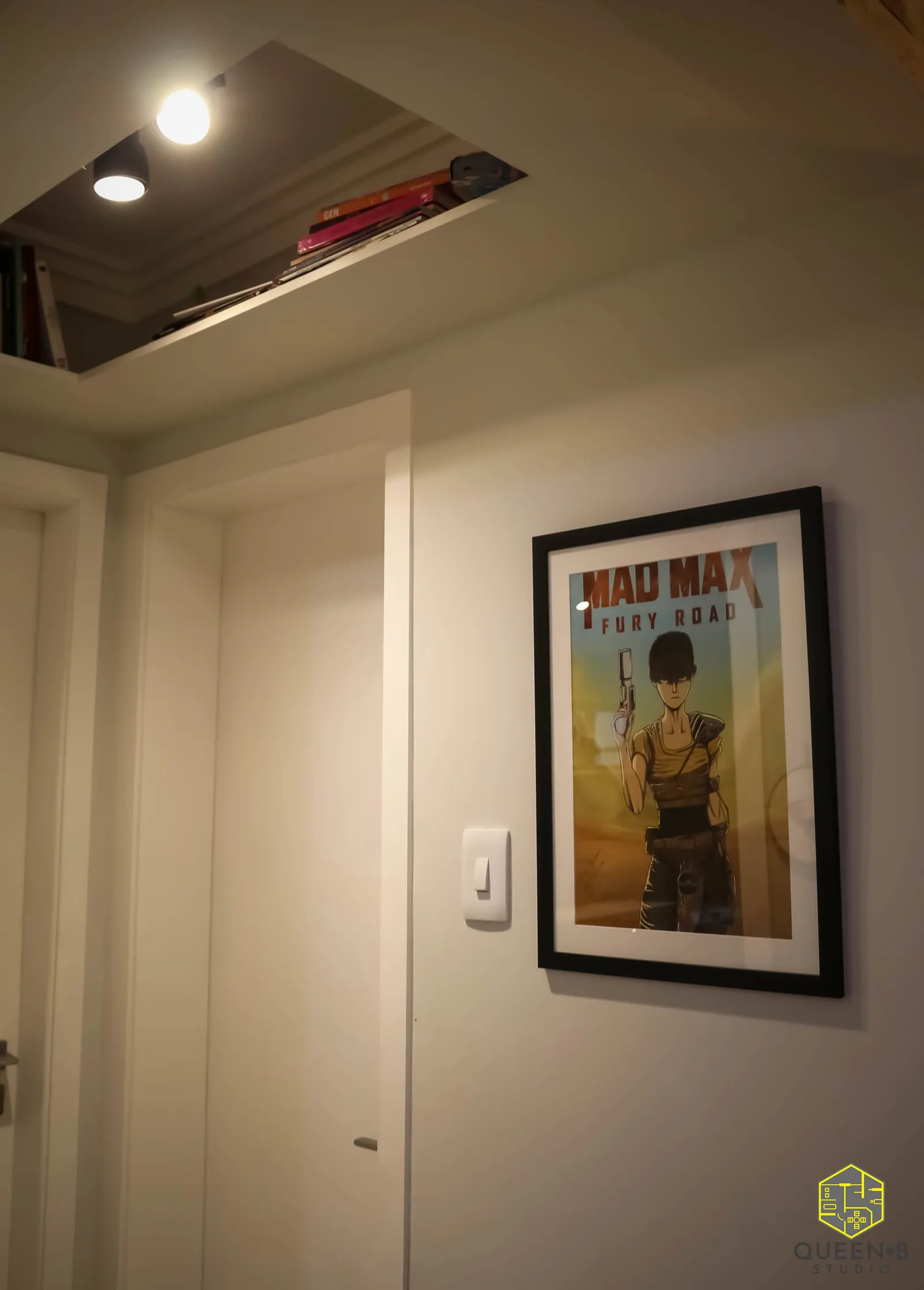 Corredor com quadro emoldurado de Mad Max Fury Road na parede branca, porta e nicho superior de teto com livros
