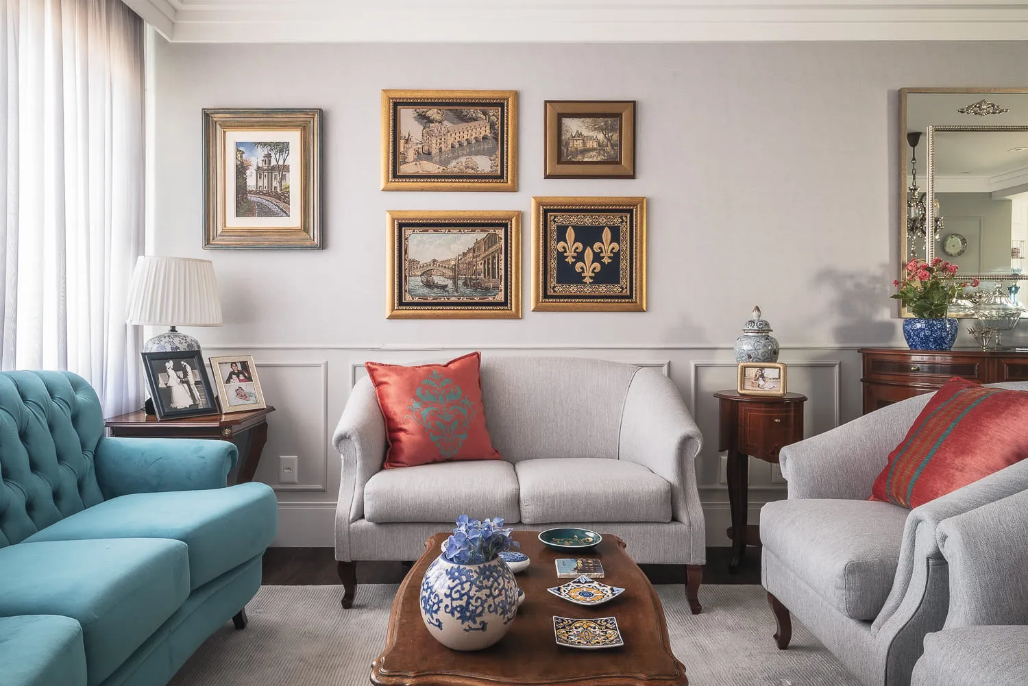 Sofá cinza com almofada vermelha bordada, poltrona azul-turquesa e parede com quadros clássicos emoldurados em dourado