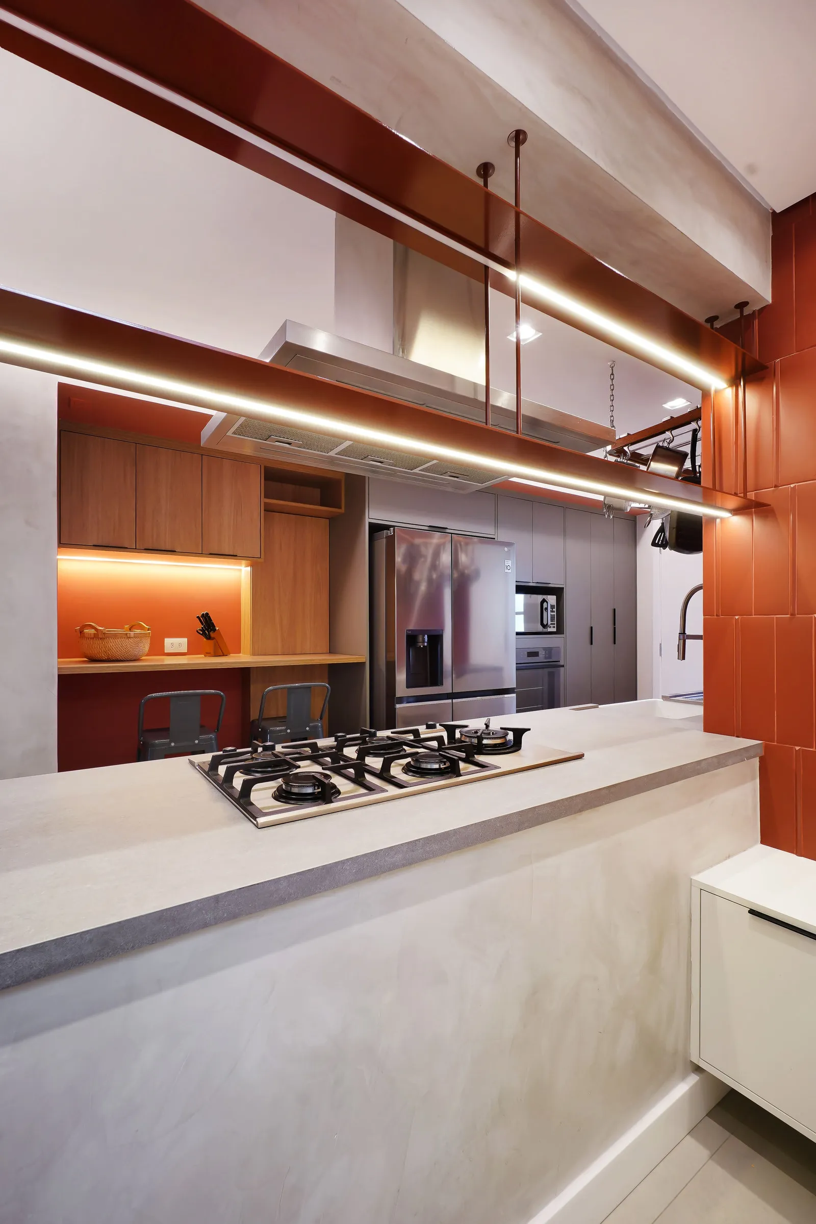 Cozinha vista pelo passa-prates, com bancada de concreto, cooktop, marcenaria amadeirada e moldura laranja