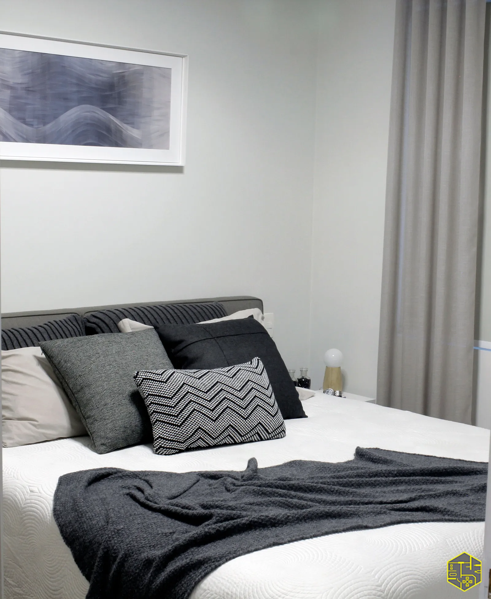 Quarto com cabeceira estofada cinza, almofadas cinza e preto, manta escura, quadro abstrato emoldurado e cortina de linho
