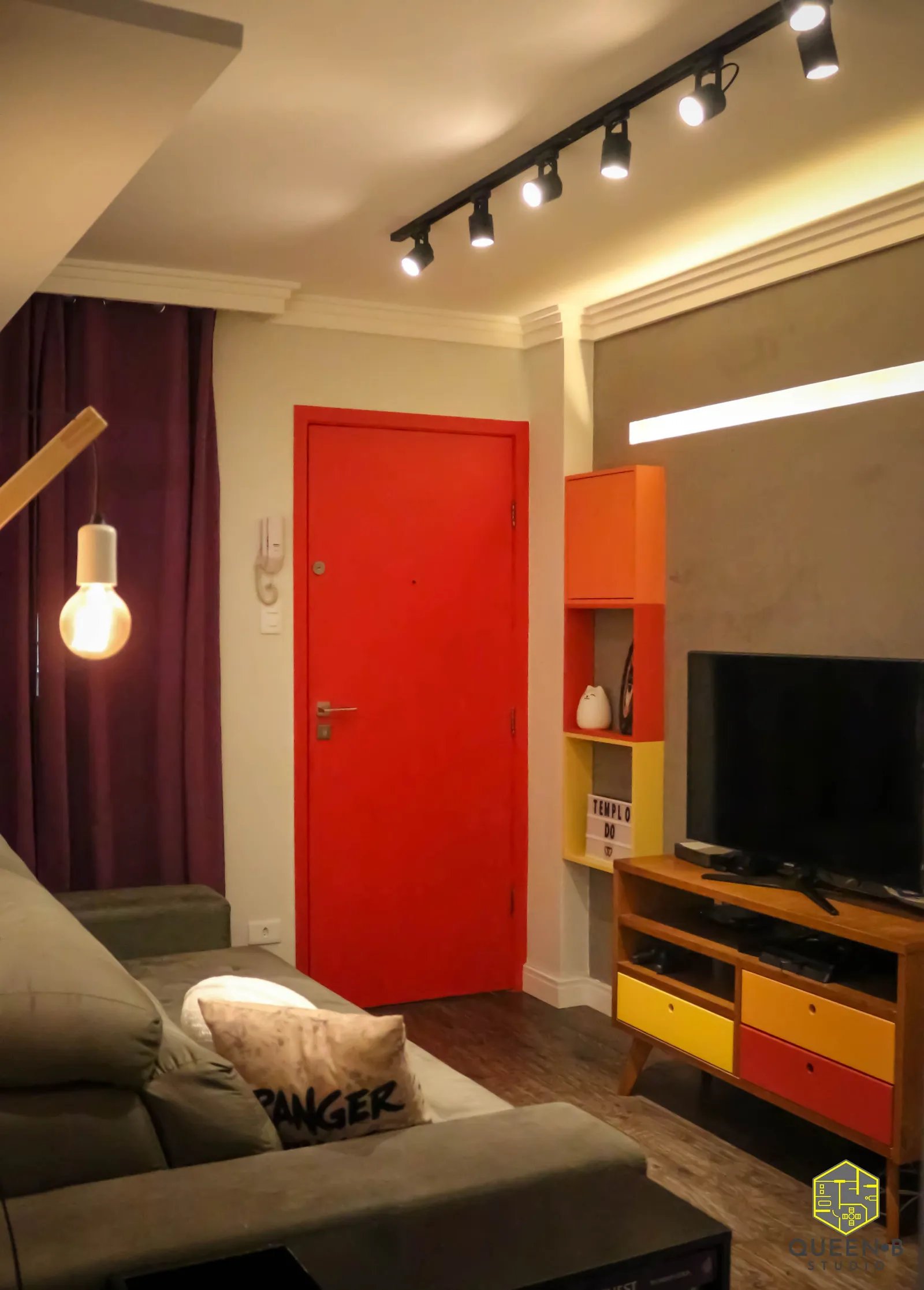 Sala com porta de entrada vermelha, sofá cinza, rack e nichos coloridos em amarelo, laranja e vermelho e trilho de spots