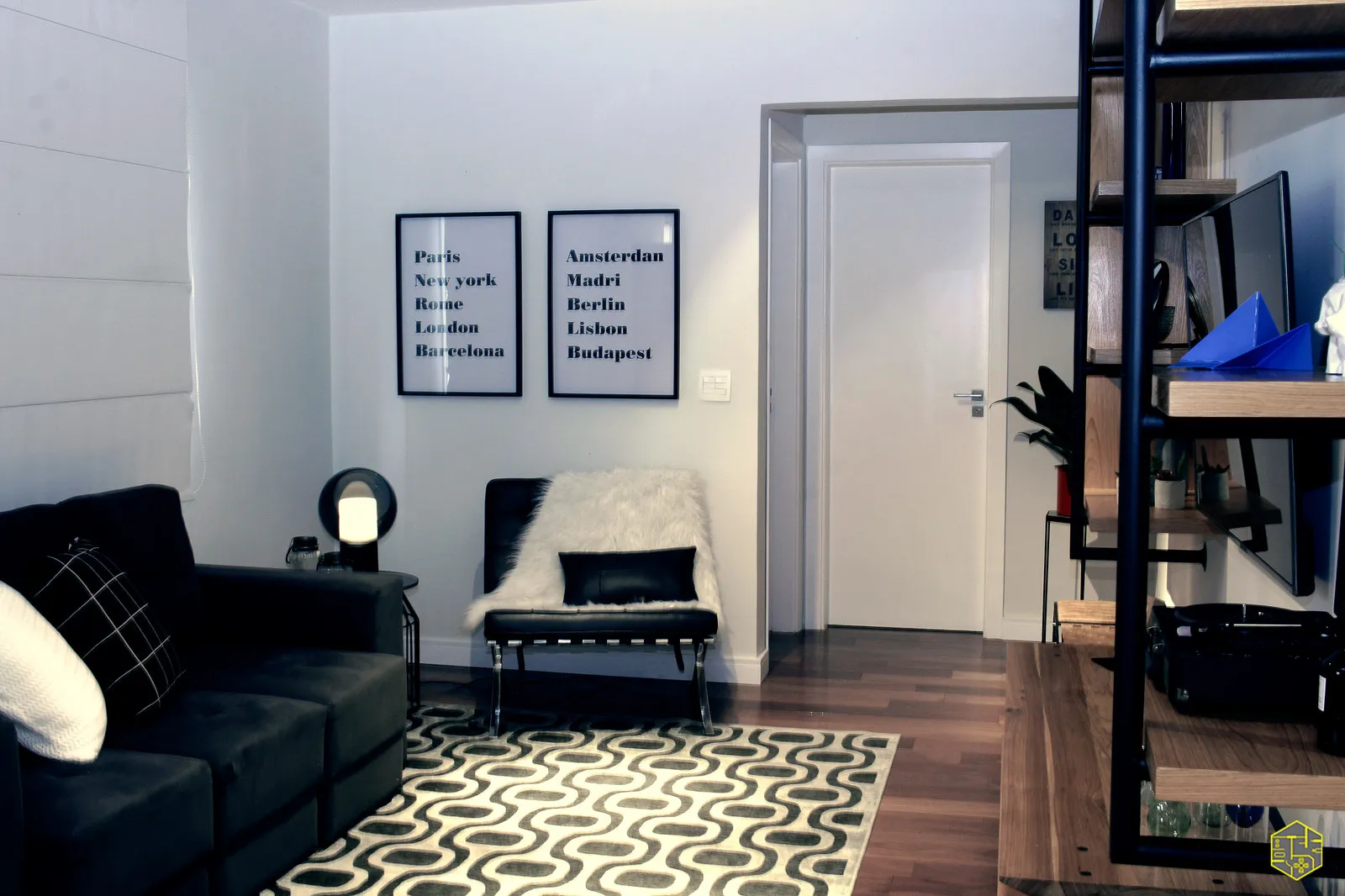 Sala com sofá preto, poltrona Barcelona com manta de pelo branco, dois quadros com nomes de cidades e tapete geométrico