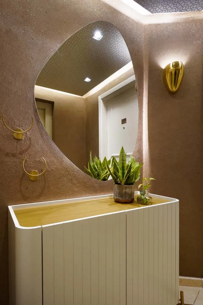 Detalhe do lavabo com espelho orgânico, arandela dourada de pétala e aparador ripado branco com tampo de madeira e plantas