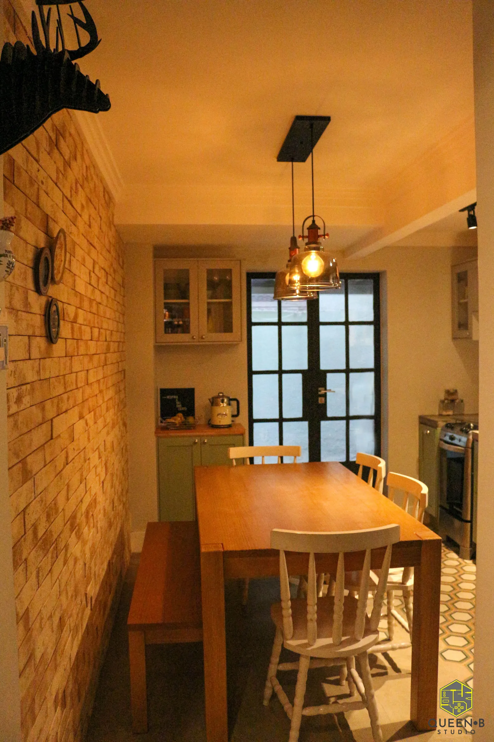 Mesa de jantar de madeira com banco e cadeiras brancas, pendentes de vidro e parede de tijolinho ao lado na cozinha