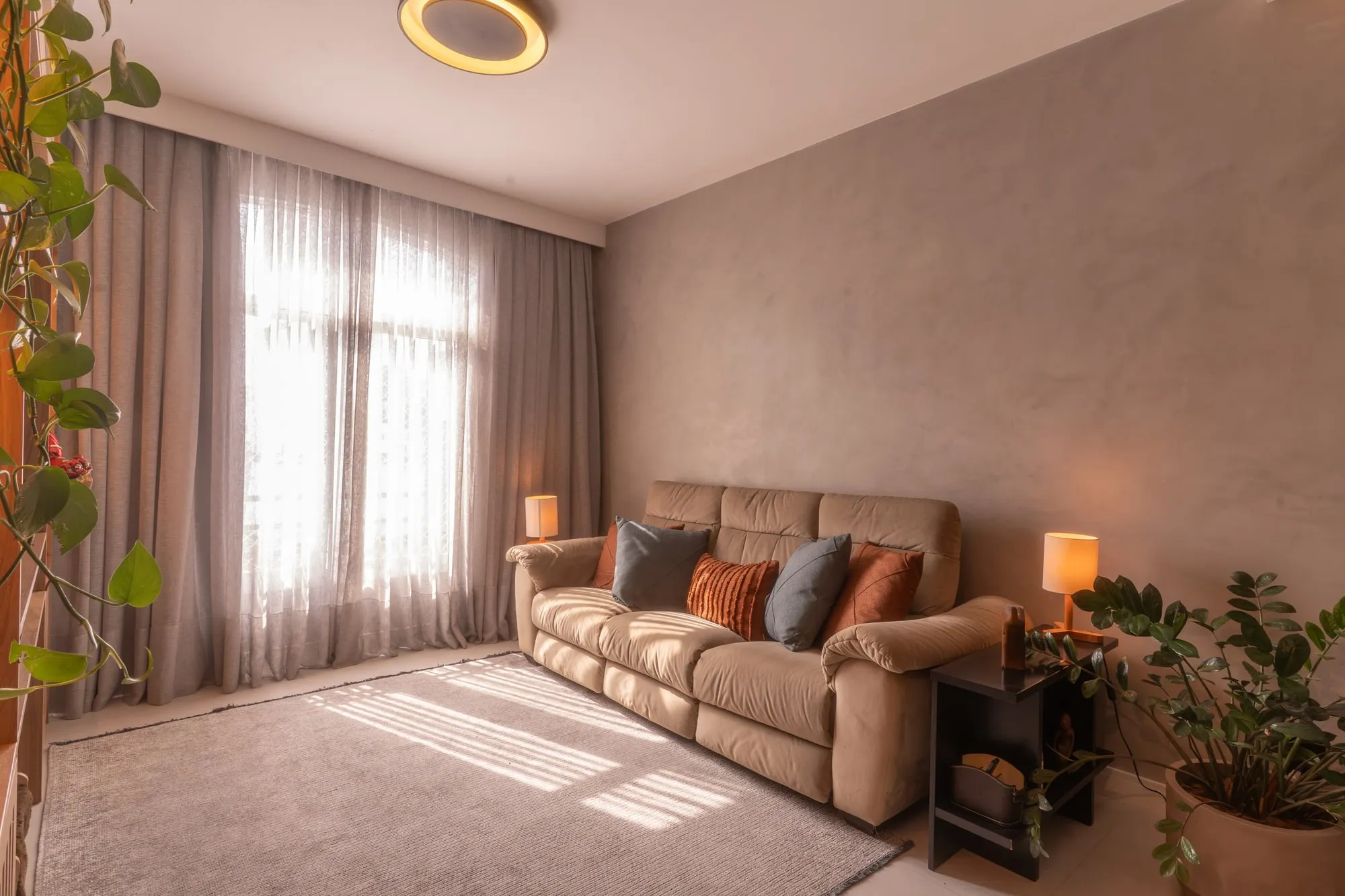 Sala de estar com sofá bege reclinável, almofadas terracota e cinza, parede de cimento queimado e plantas