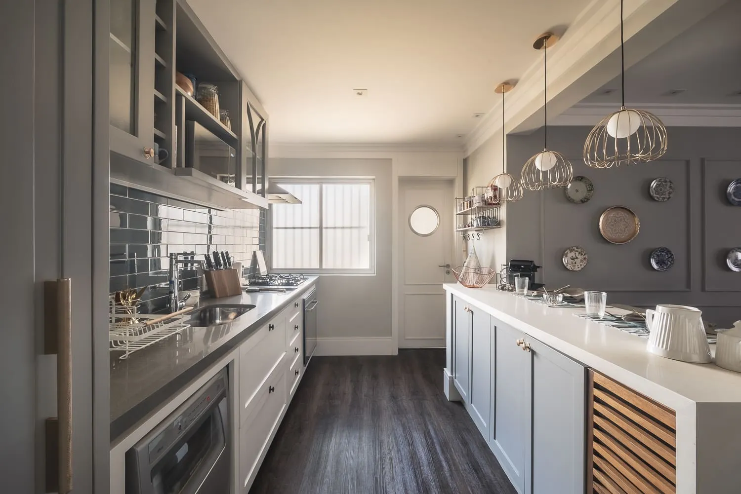 Cozinha em corredor com marcenaria cinza e branca, backsplash azul escuro, pendentes dourados e parede de pratos decorativos