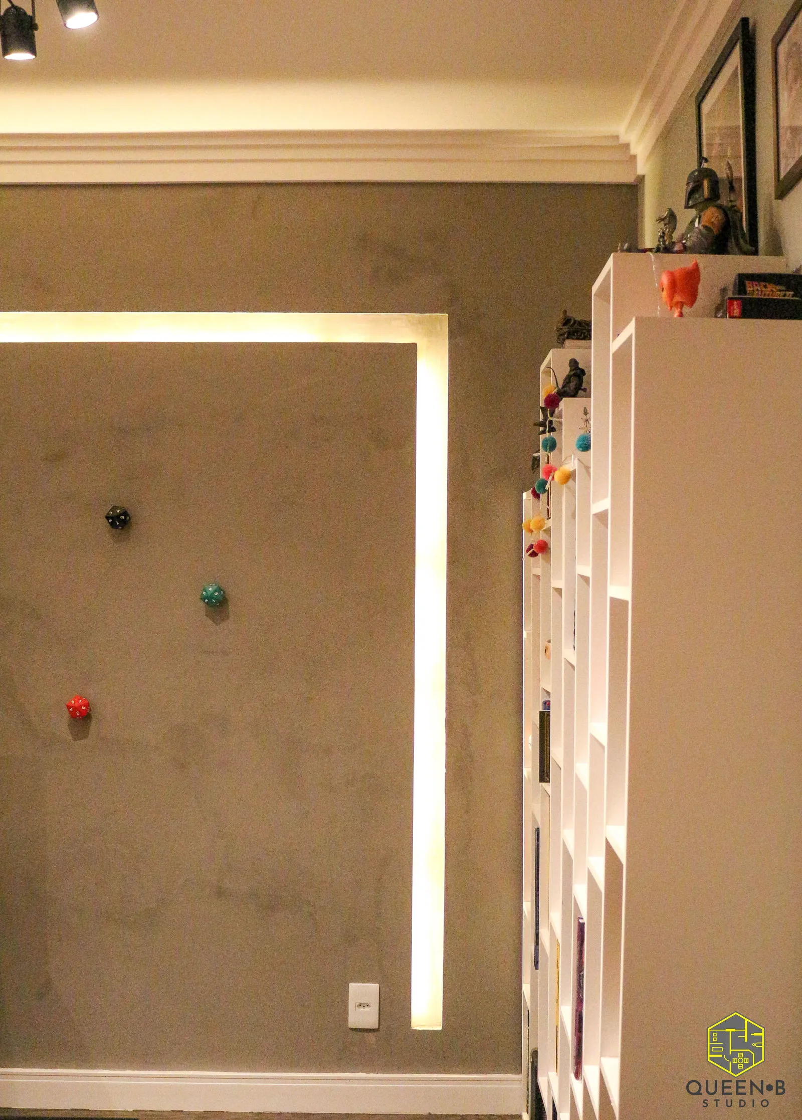 Parede de cimento queimado com dados de RPG aplicados e nicho de LED, ao lado de estantes brancas com livros e colecionáveis