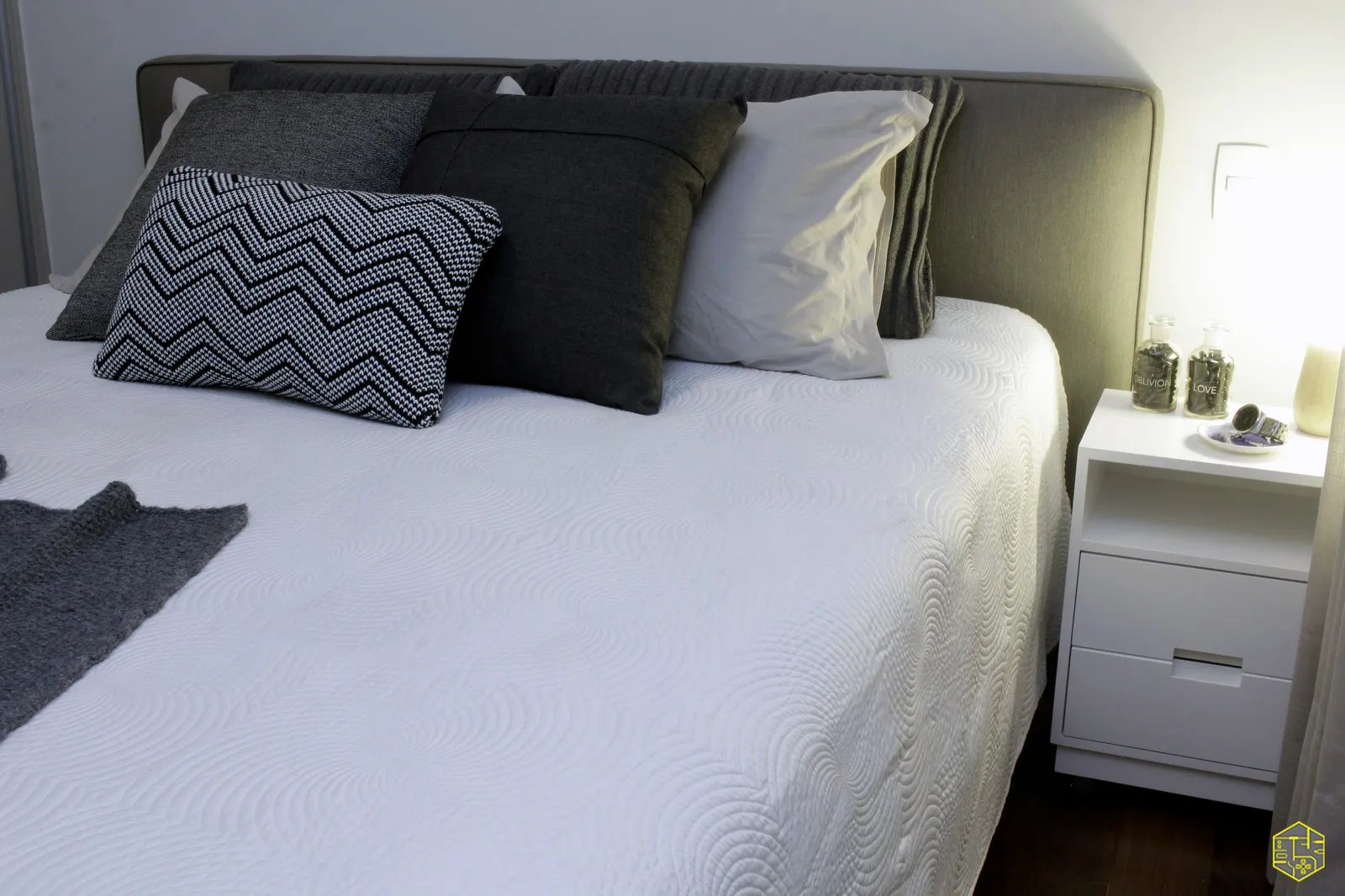 Detalhe do quarto com cama de colcha branca, almofadas cinza e estampada e criado-mudo branco com frascos de vidro