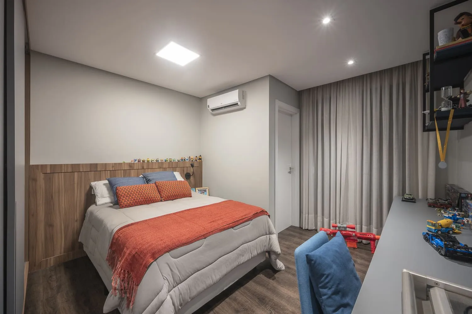 Quarto infantil com cama de colcha cinza e mantas laranja, cabeceira amadeirada e bancada com brinquedos