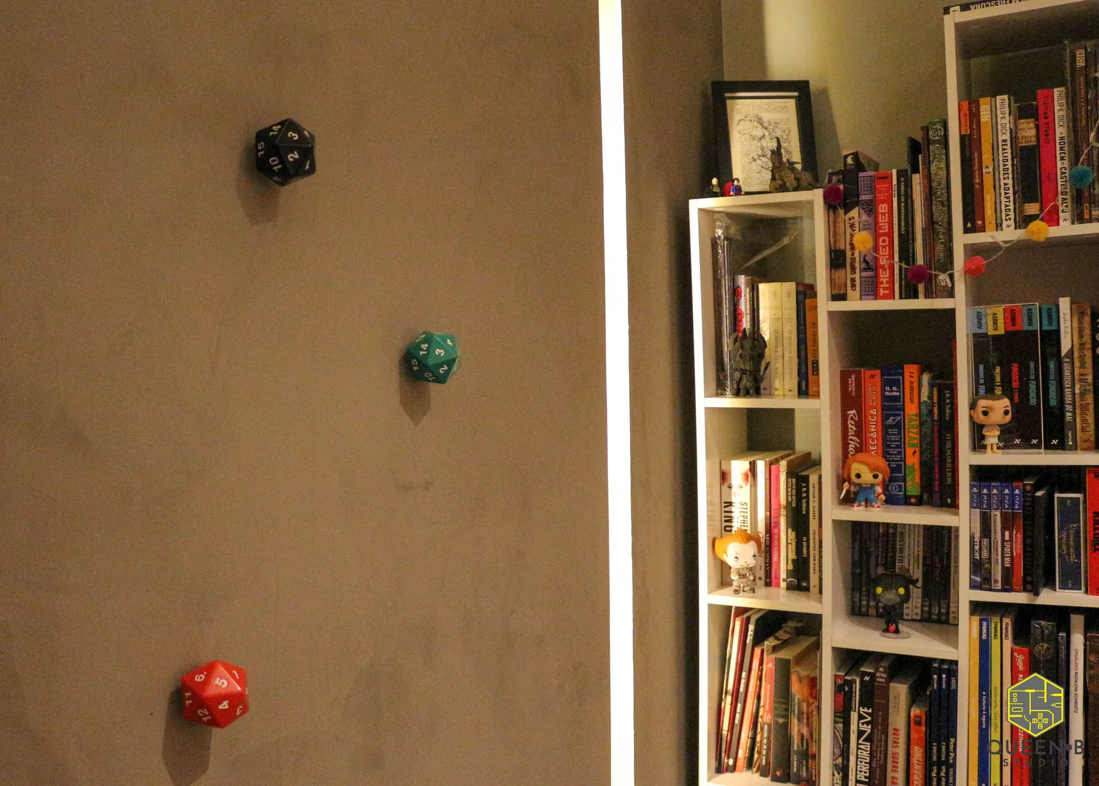 Parede de cimento queimado com três dados de RPG preto, verde e vermelho, ao lado de estante branca cheia de livros e bonecos