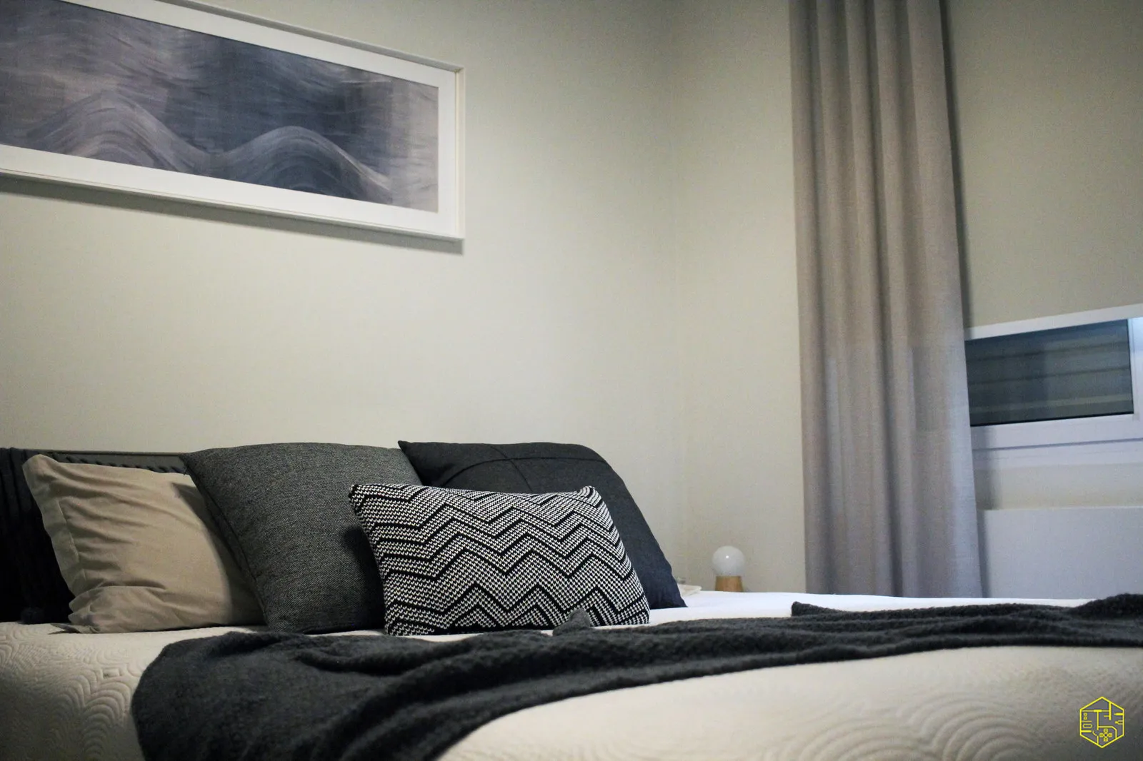 Quarto em ângulo baixo com almofadas cinza e estampada, manta escura, quadro panorâmico abstrato e cortina cinza