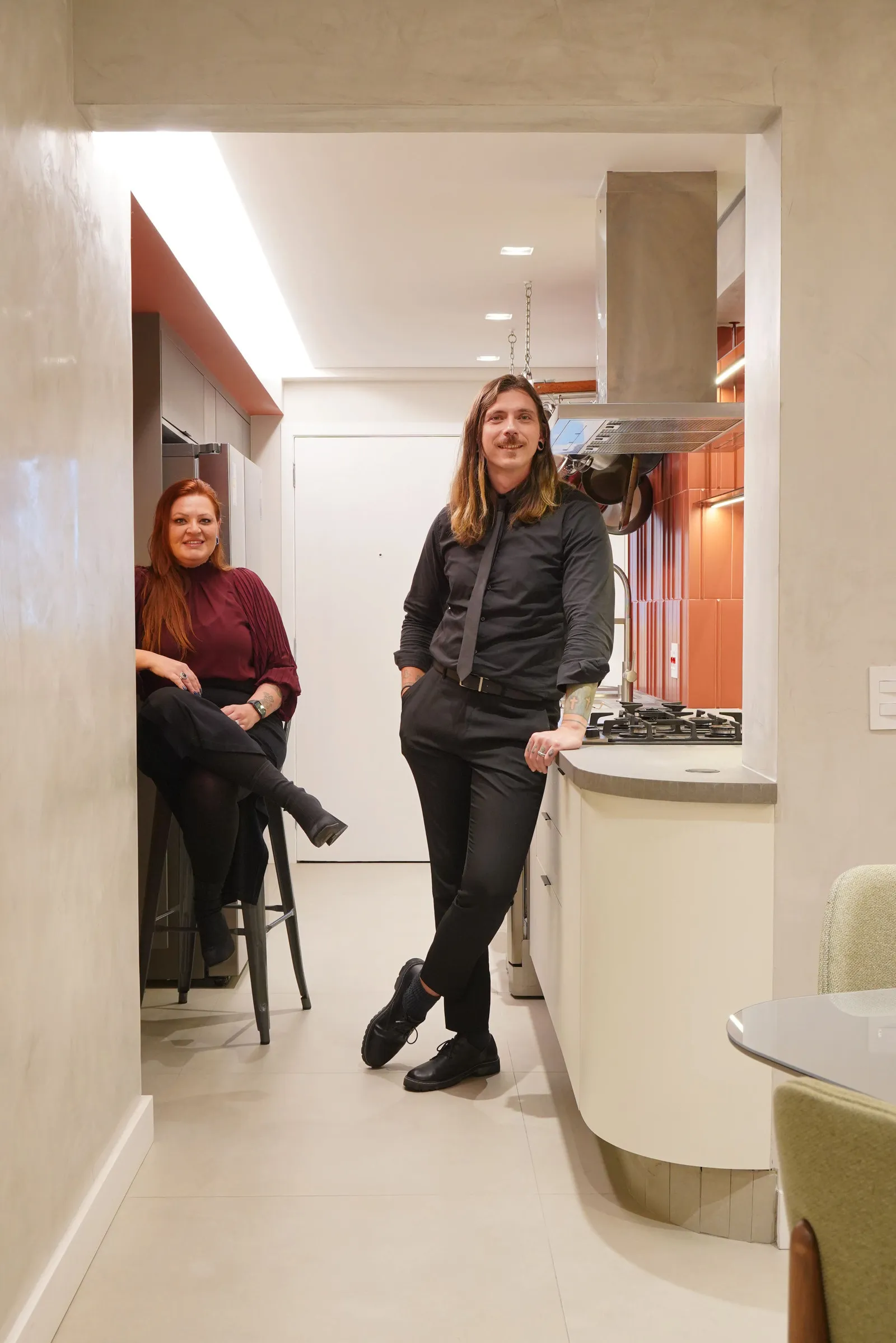 Casal na entrada da cozinha, ele em pé junto à bancada e ela sentada na banqueta ao fundo