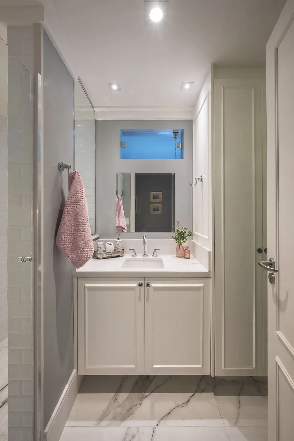 Banheiro branco com bancada de mármore, gabinete clássico off-white, espelho, toalha rosa e piso de mármore