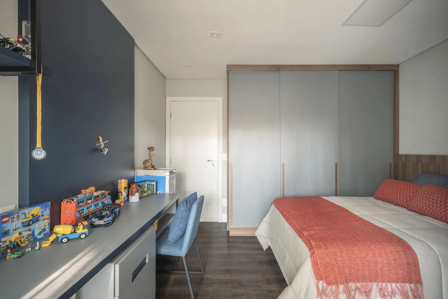 Quarto infantil com parede azul-marinho, bancada de estudos com brinquedos, guarda-roupa cinza e cama ao lado